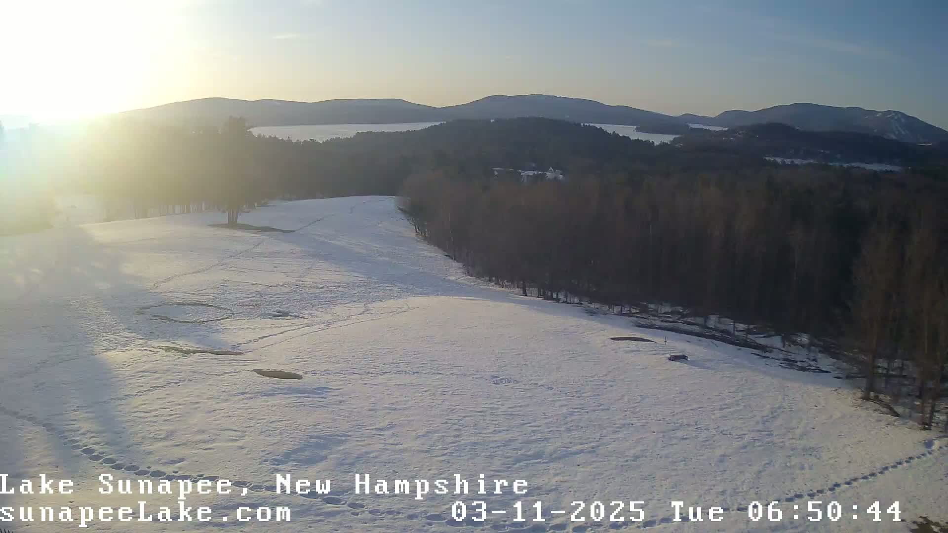 A snow-covered field with a line of trees bordering a lake in the distance, under a bright, sunny sky.
