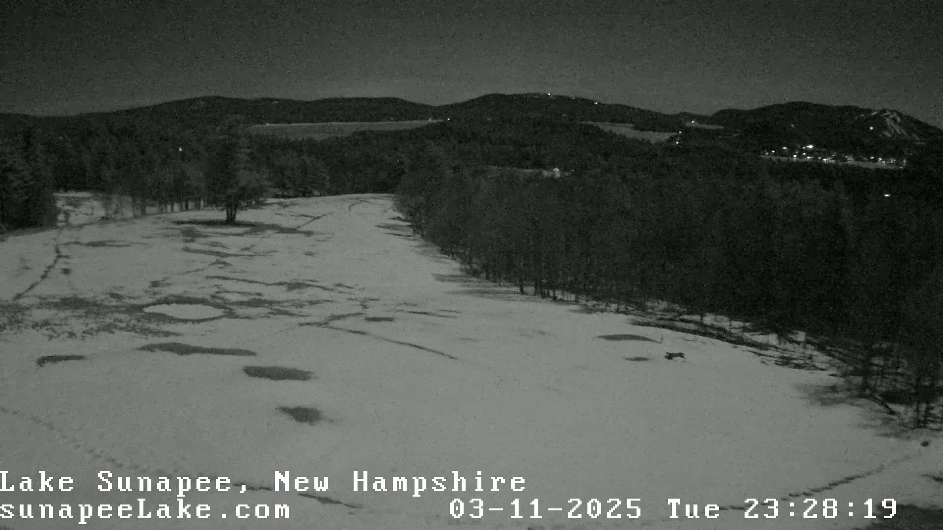 A snow-covered field at night, bordered by dark trees and distant hills with a few lights visible.