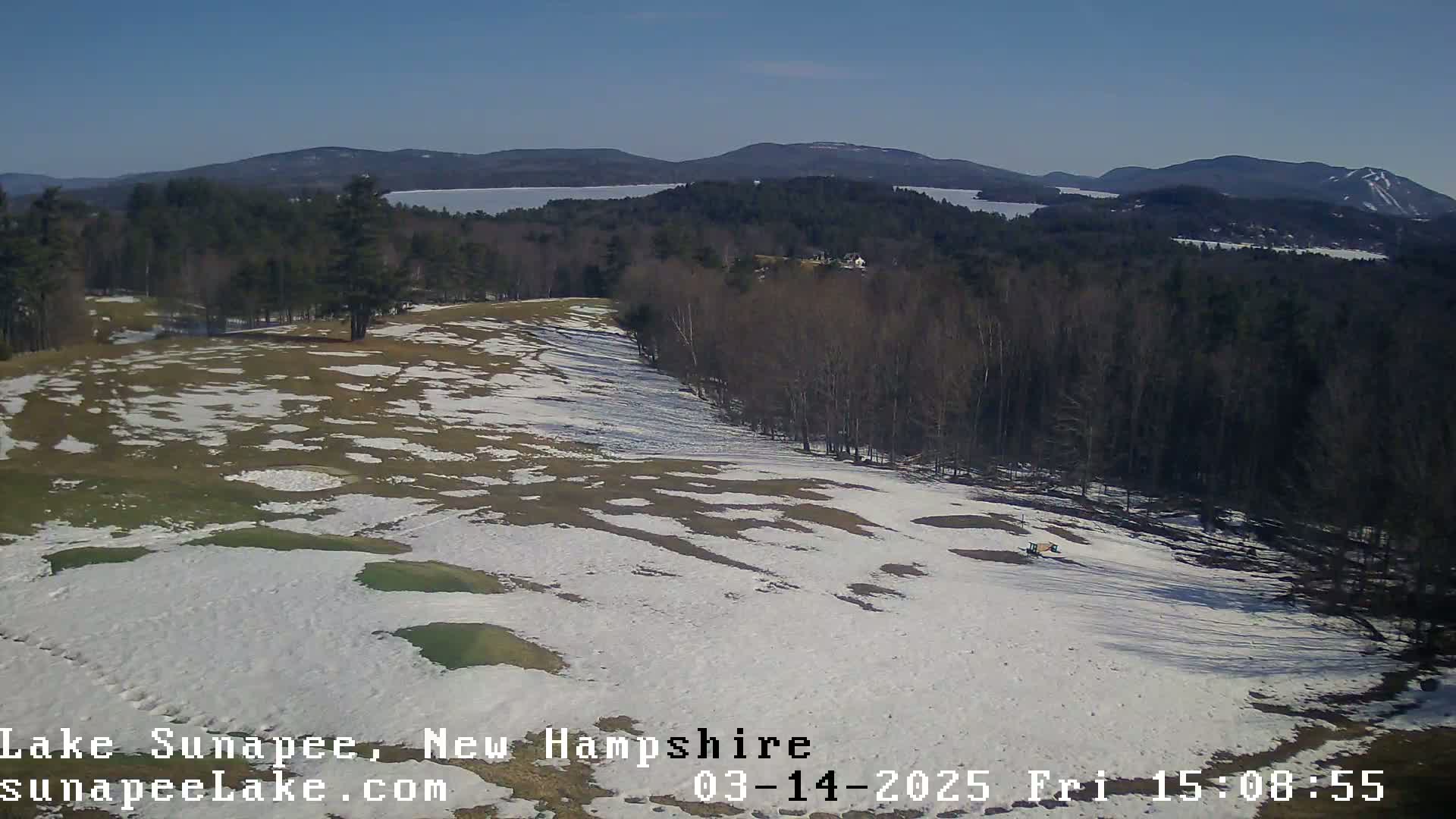 A partially snow-covered golf course overlooks a lake and distant mountains under a clear blue sky.
