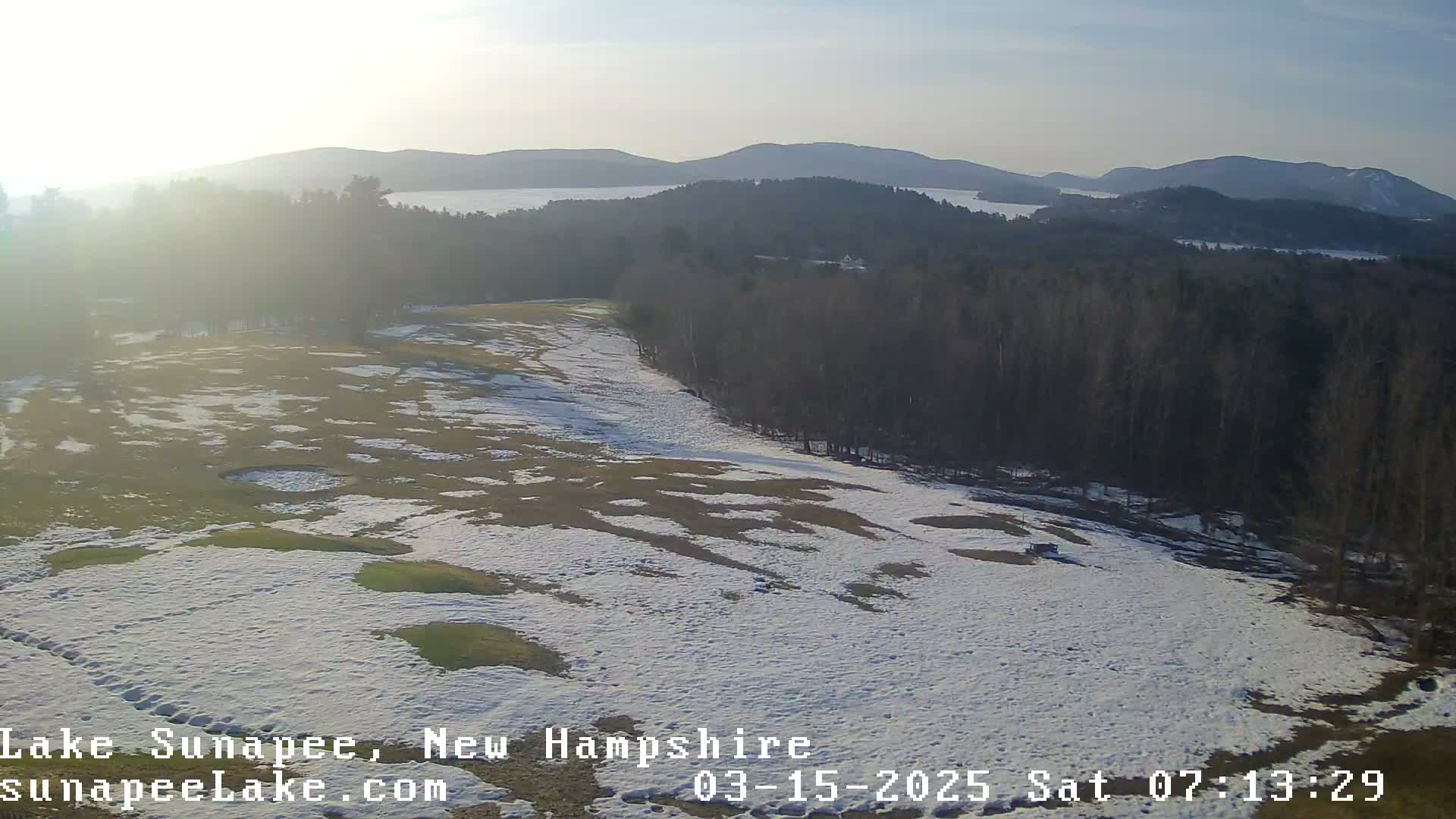 A partially snow-covered grassy area slopes down towards a line of trees and a distant lake and mountains under a bright, hazy sky.
