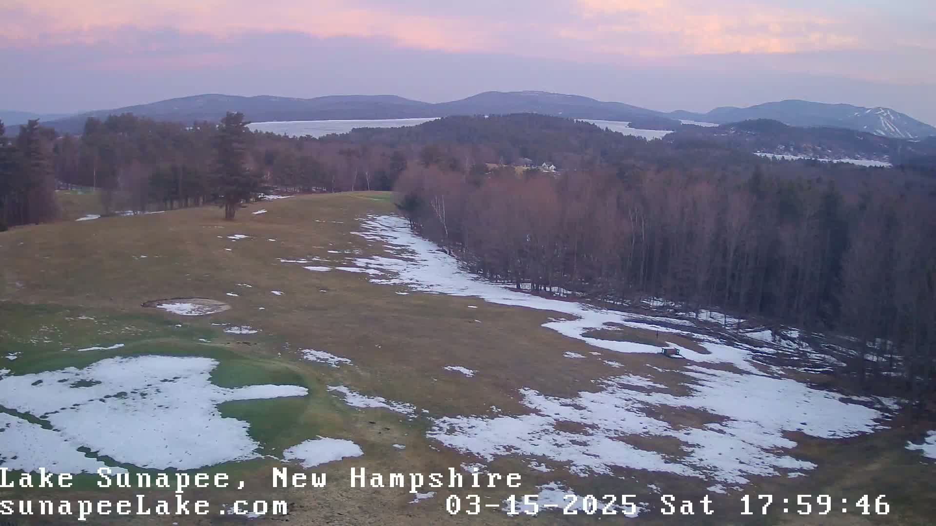 A partially snow-covered golf course overlooks a lake and distant mountains under a soft pink sunset.