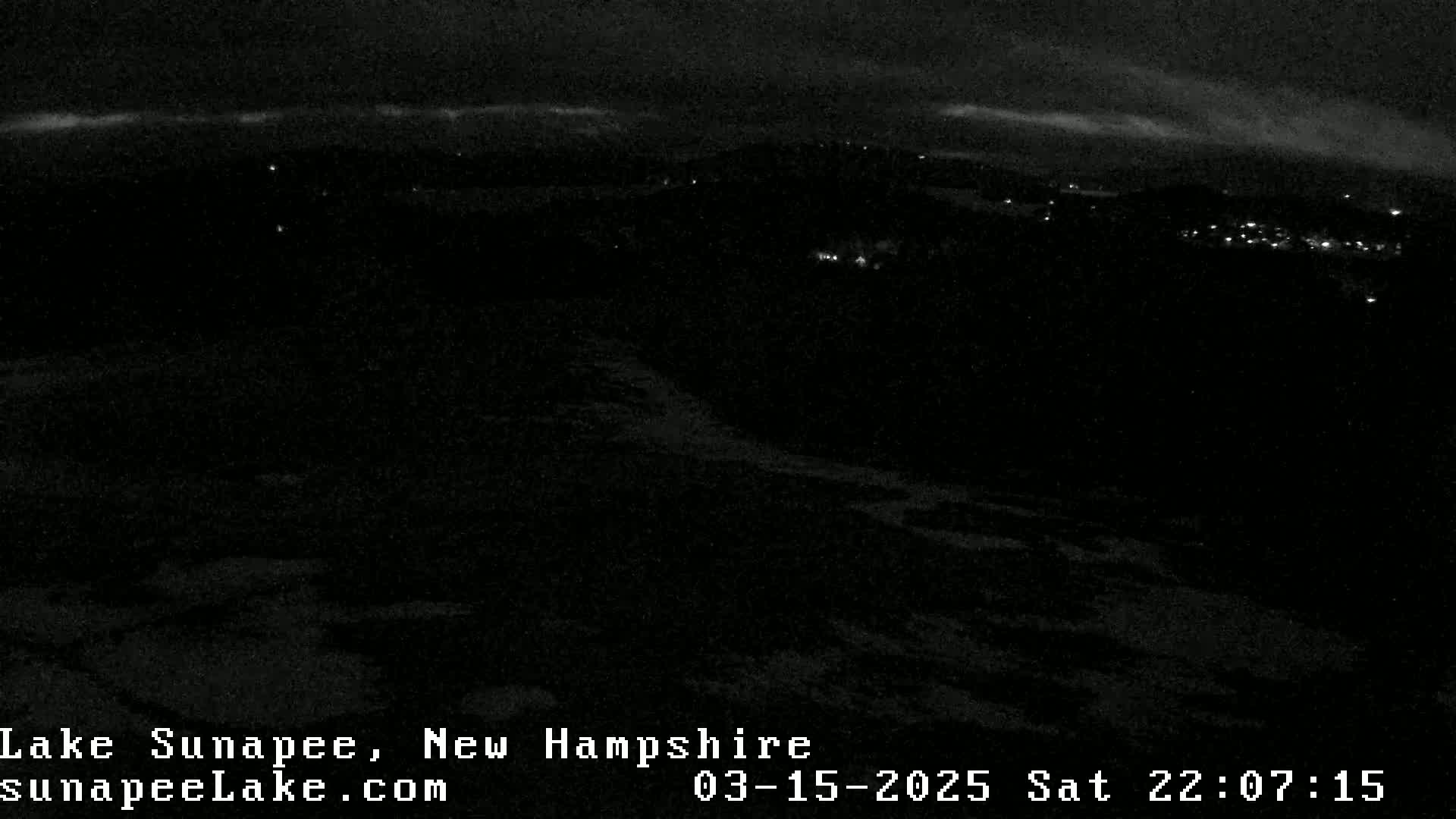 A nighttime, low-light view of a dark landscape with a few scattered lights visible in the distance under a mostly cloudy sky.