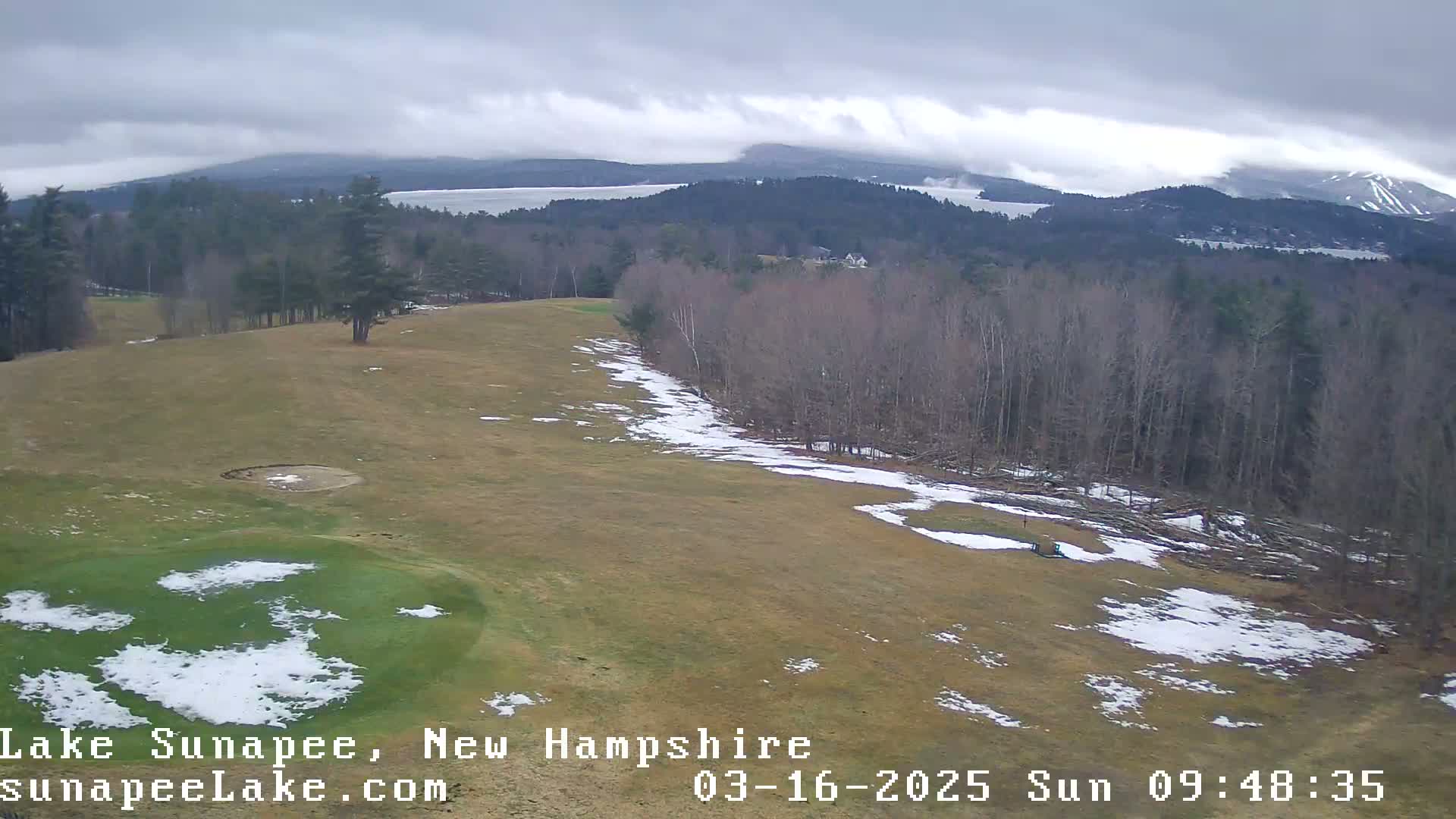 A partly snow-covered golf course overlooks a lake and distant mountains under an overcast sky.