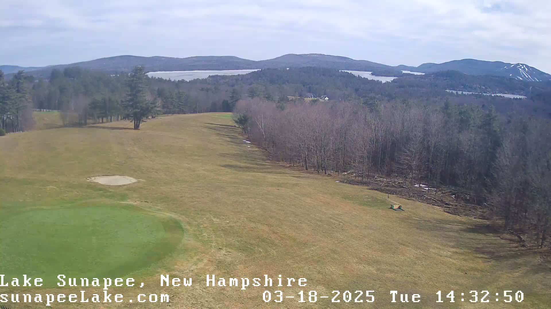 A golf course green and fairway are shown, overlooking a lake and distant mountains under a mostly clear sky.