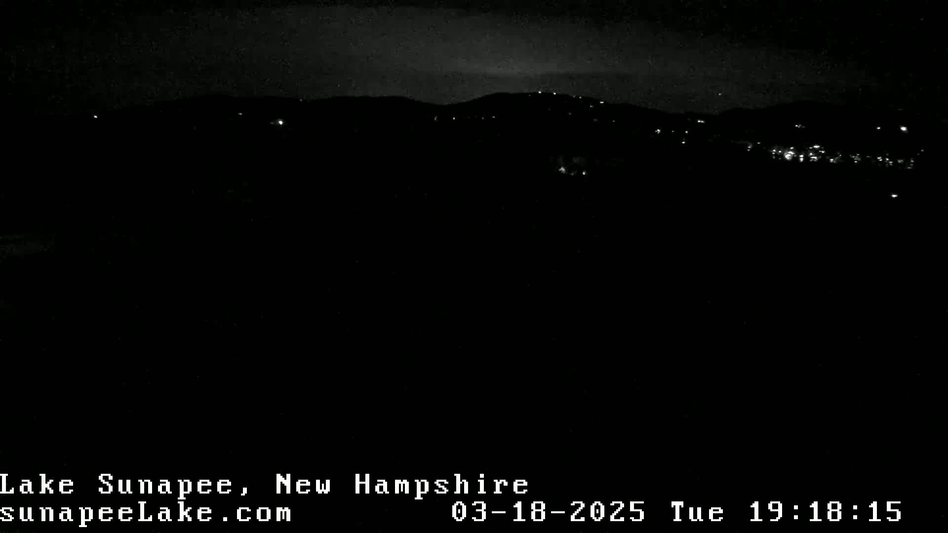 A nighttime long shot of a dark landscape with a few distant lights under a mostly clear sky.
