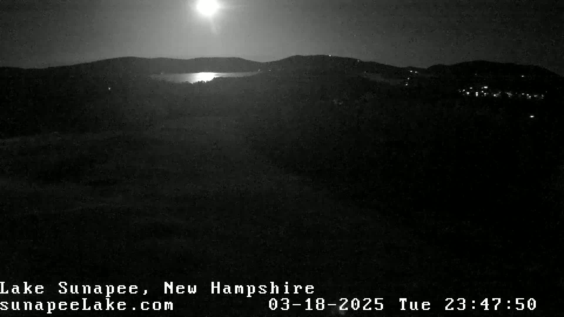 A nighttime view of a moonlit lake surrounded by dark hills and a few distant lights.