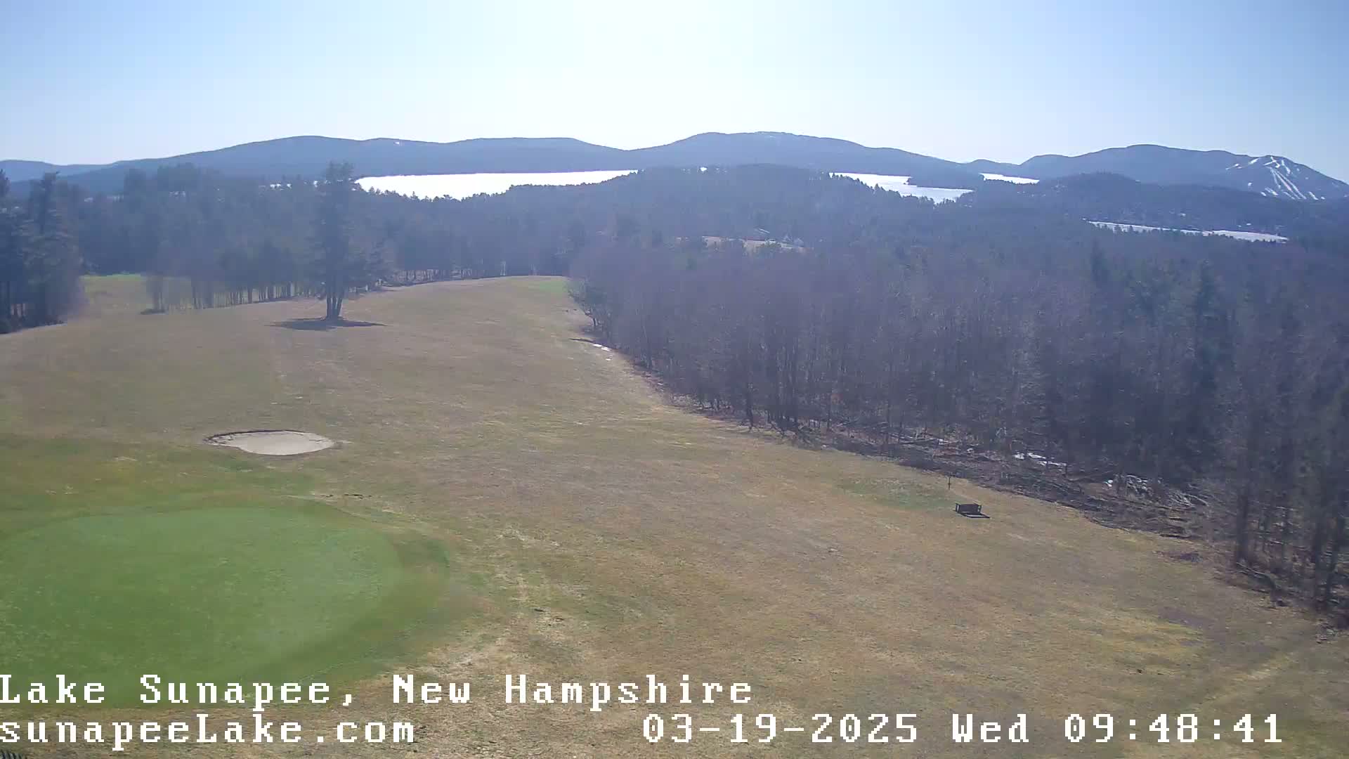 A high-angle view of a golf course on a sunny day, overlooking a lake and distant mountains with some snow on the higher peaks.