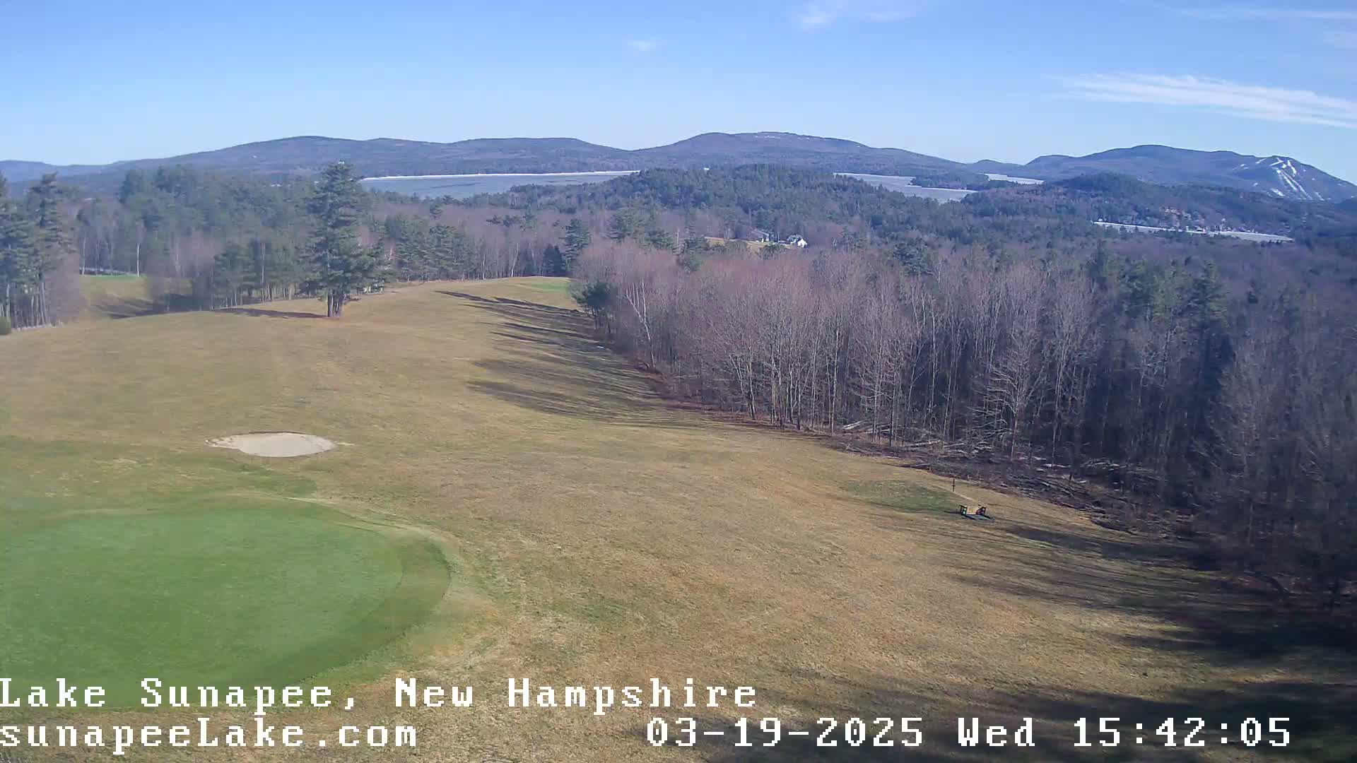 A sunny day reveals a golf course, bordered by leafless trees, overlooking a distant lake and mountains.