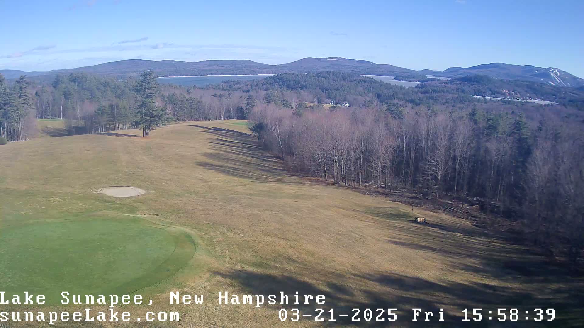 An elevated view of a golf course with a green and sand trap, overlooking a lake and distant mountains under a clear, sunny sky.