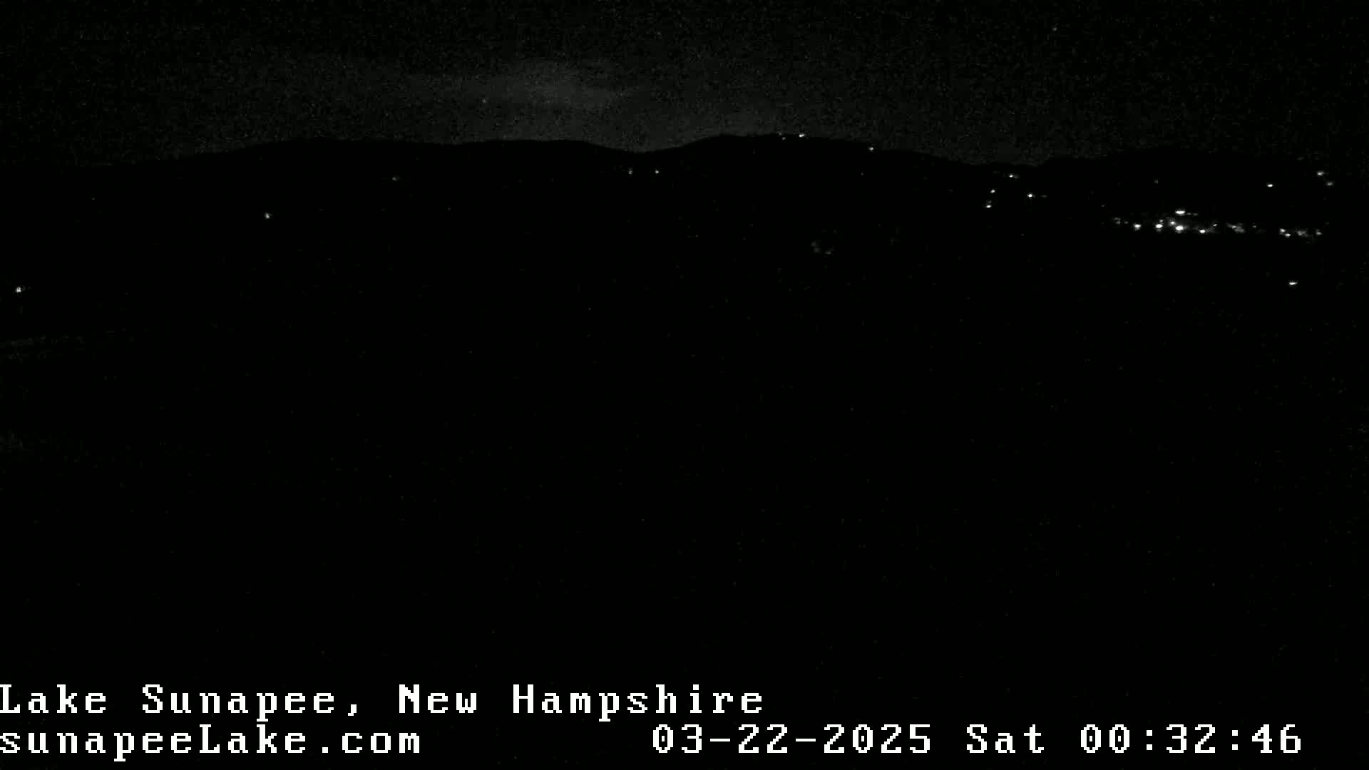 A nighttime long shot of a dark landscape with faintly visible distant lights under a mostly clear sky.