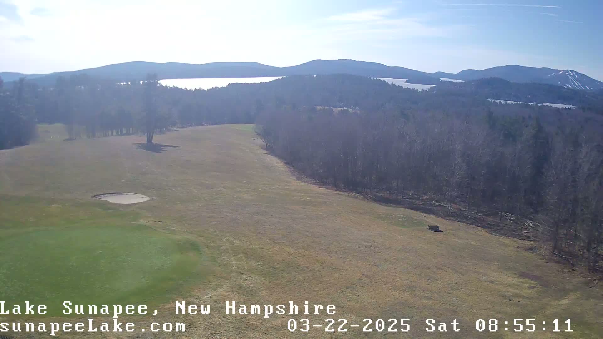 A high-angle view shows a golf course with a sand trap, bordered by a forest, overlooking a lake and distant mountains under a clear, sunny sky.