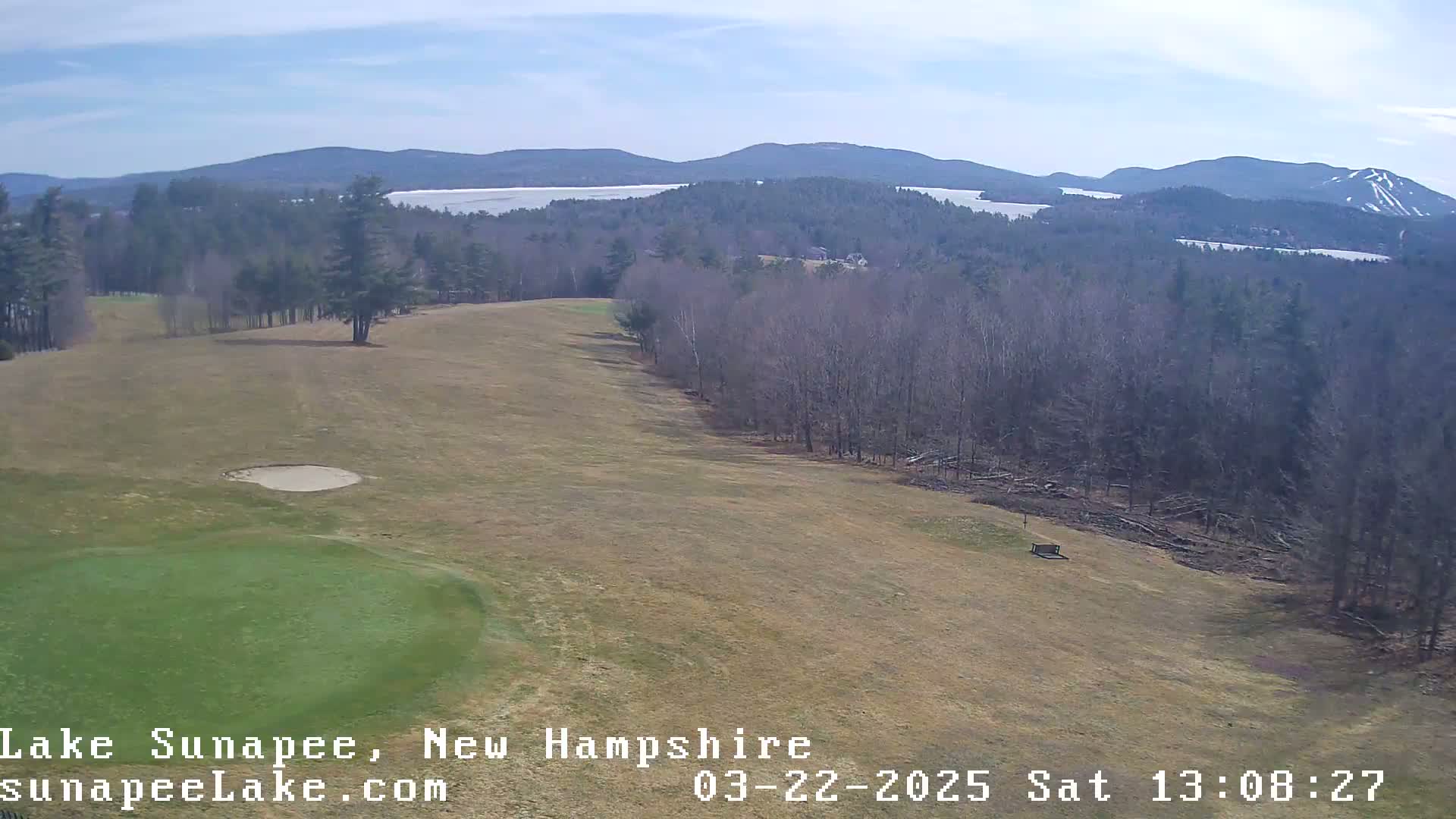 A golf course fairway slopes down towards a lake and distant mountains under a mostly clear sky.