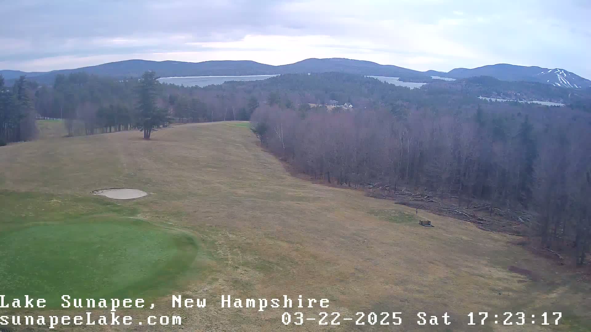 An elevated view of a golf course green and sand trap, overlooking a mostly barren landscape with distant mountains and a lake under an overcast sky.