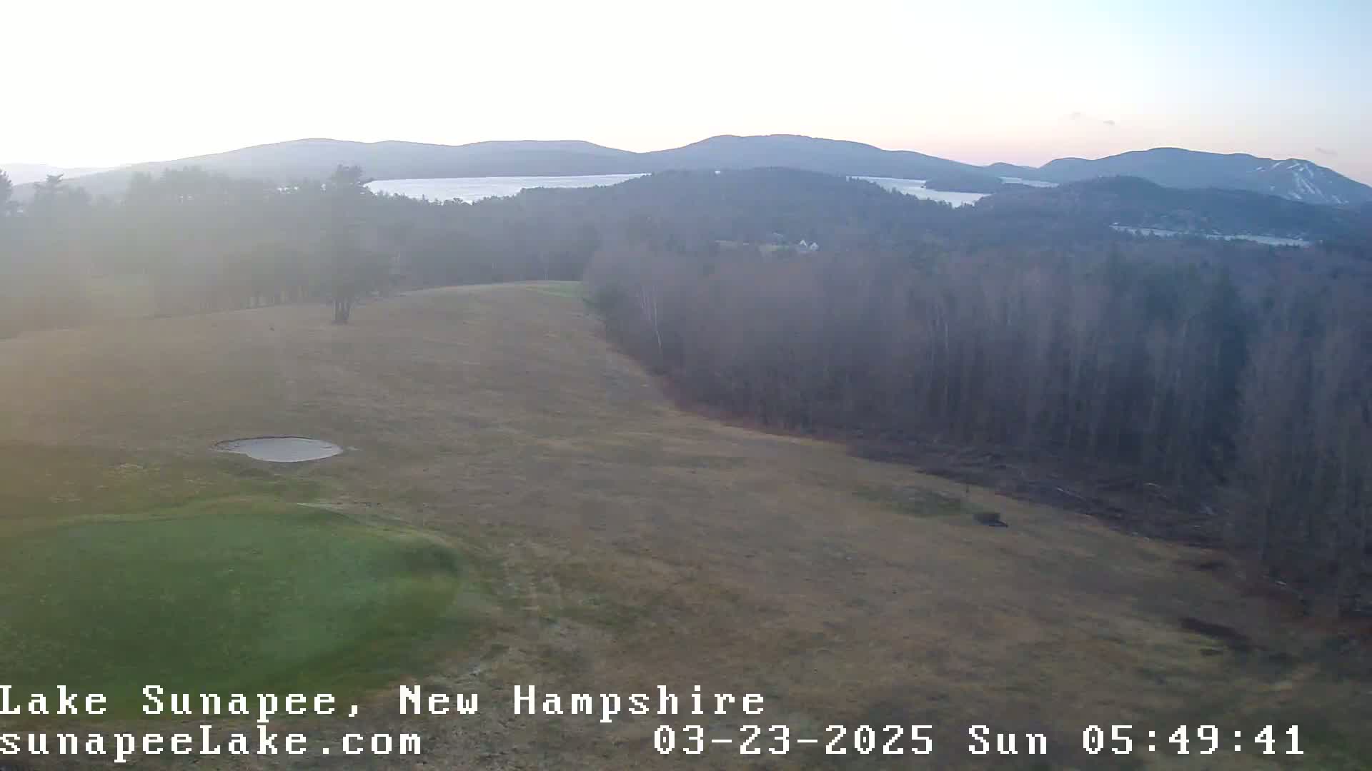 A hazy sunrise reveals a golf course overlooking a lake and distant mountains.