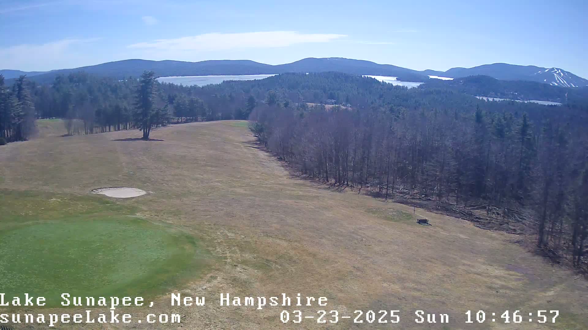 A high-angle view shows a golf course with a green and sand trap, bordered by a line of leafless trees, and a lake and mountains in the background under a clear, sunny sky.
