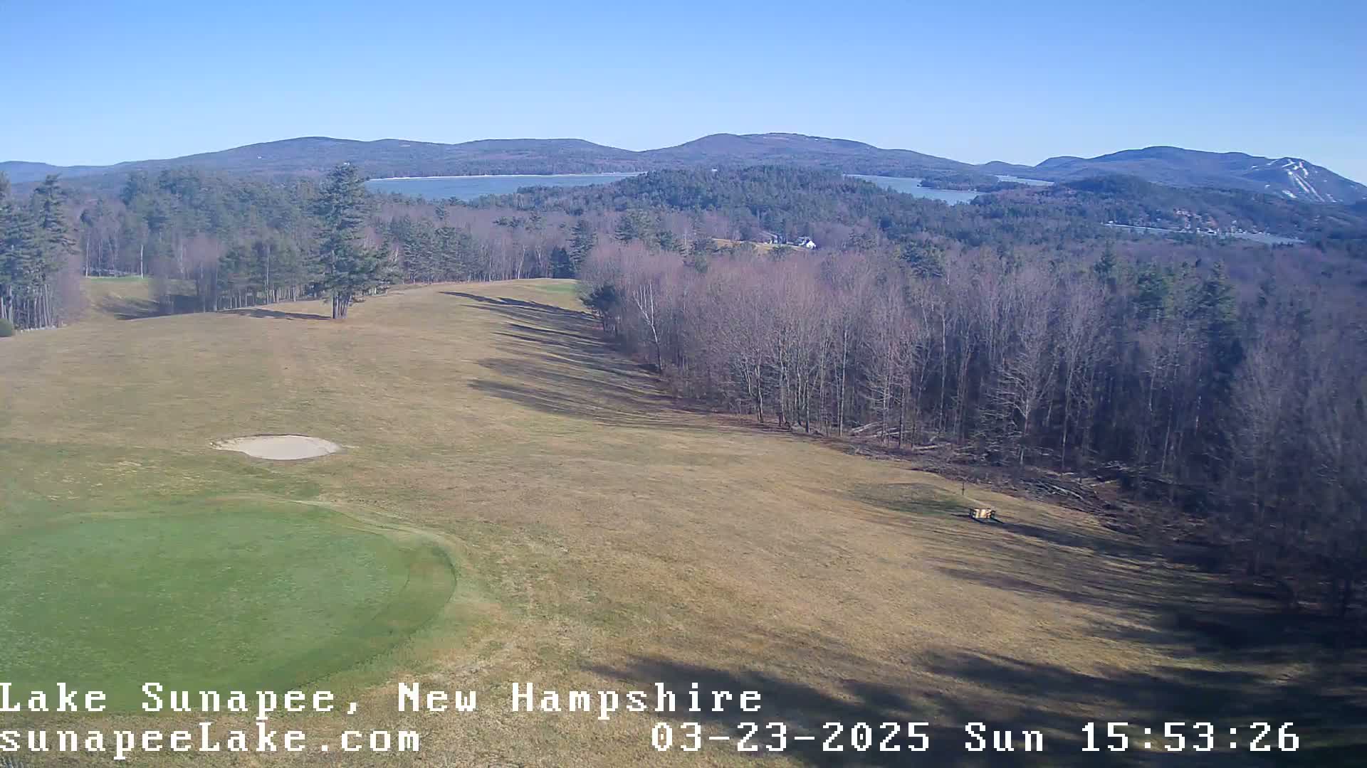 A sunny view of a golf course with a sand trap and putting green, overlooking a lake and distant mountains.