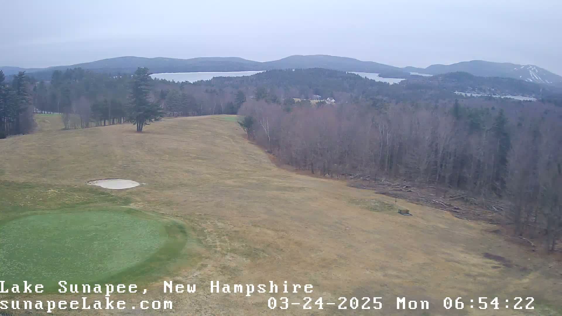An elevated view of a golf course green and fairway, overlooking a lake and distant mountains under an overcast sky.
