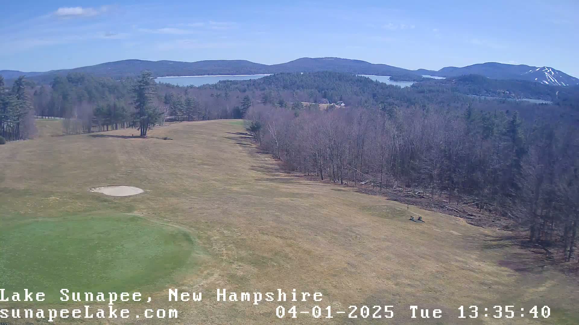 A high-angle view shows a mostly-bare golf course with a green and sand trap, overlooking a lake and distant mountains under a clear blue sky.