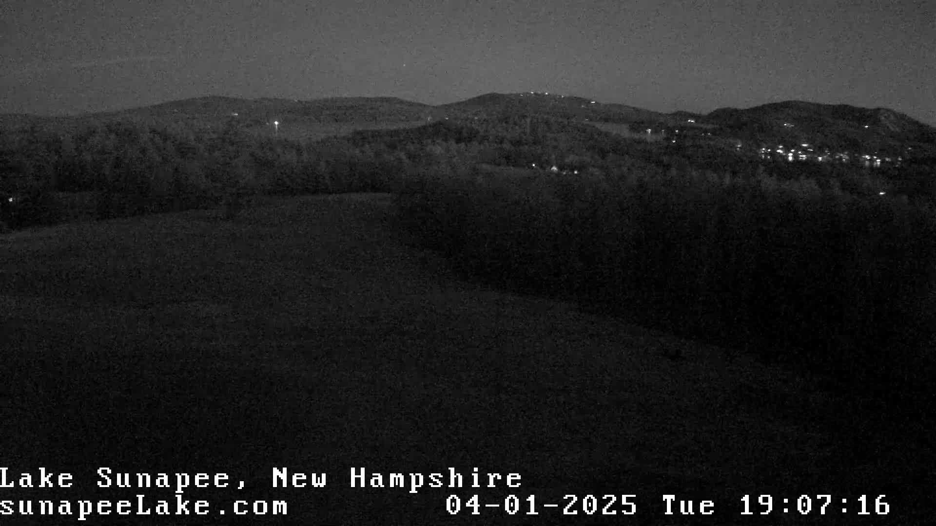 A nighttime view of a dark landscape with distant lights and hills under a clear, dark sky.