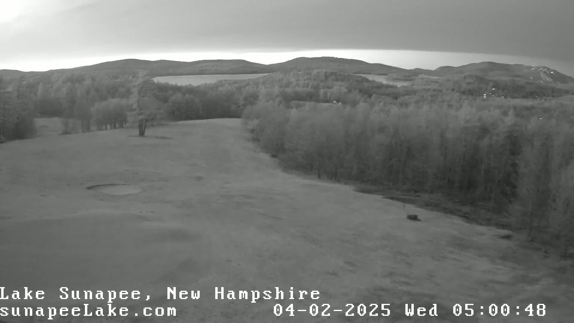 A grayscale image shows a golf course with a sand trap, bordered by trees and overlooking a lake and distant mountains under an overcast sky.