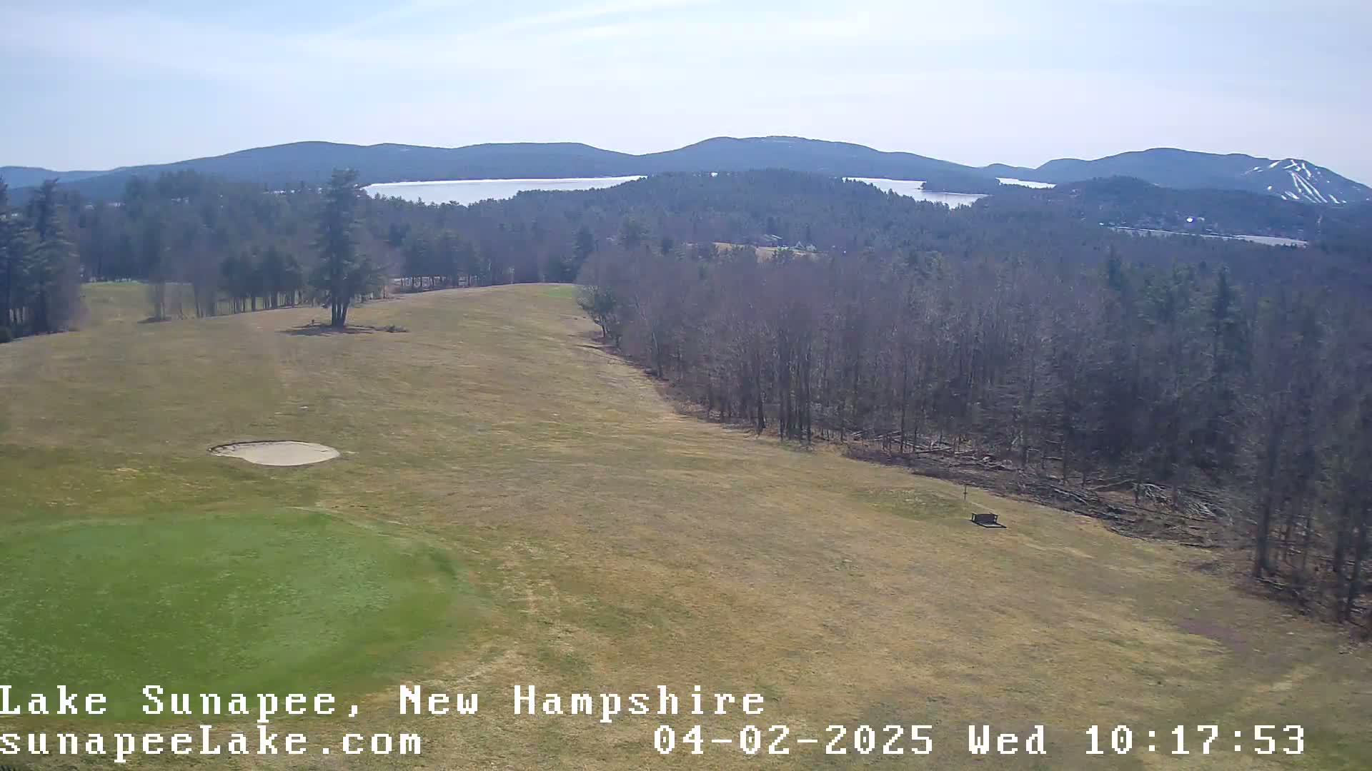 A mostly clear day reveals a golf course and a distant lake nestled among bare, winter trees and mountains, with a hint of snow on a distant peak.