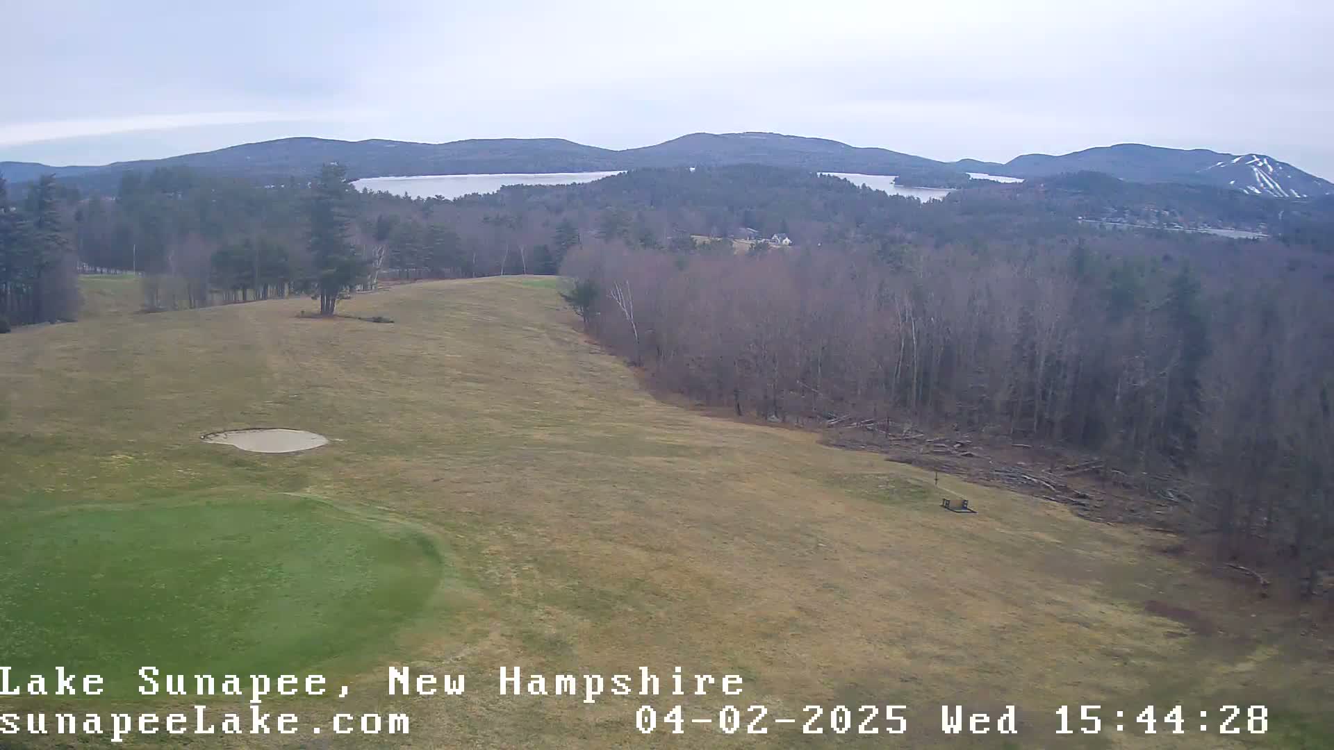 A golf course with a sand trap and green, overlooking a lake and distant mountains under an overcast sky.
