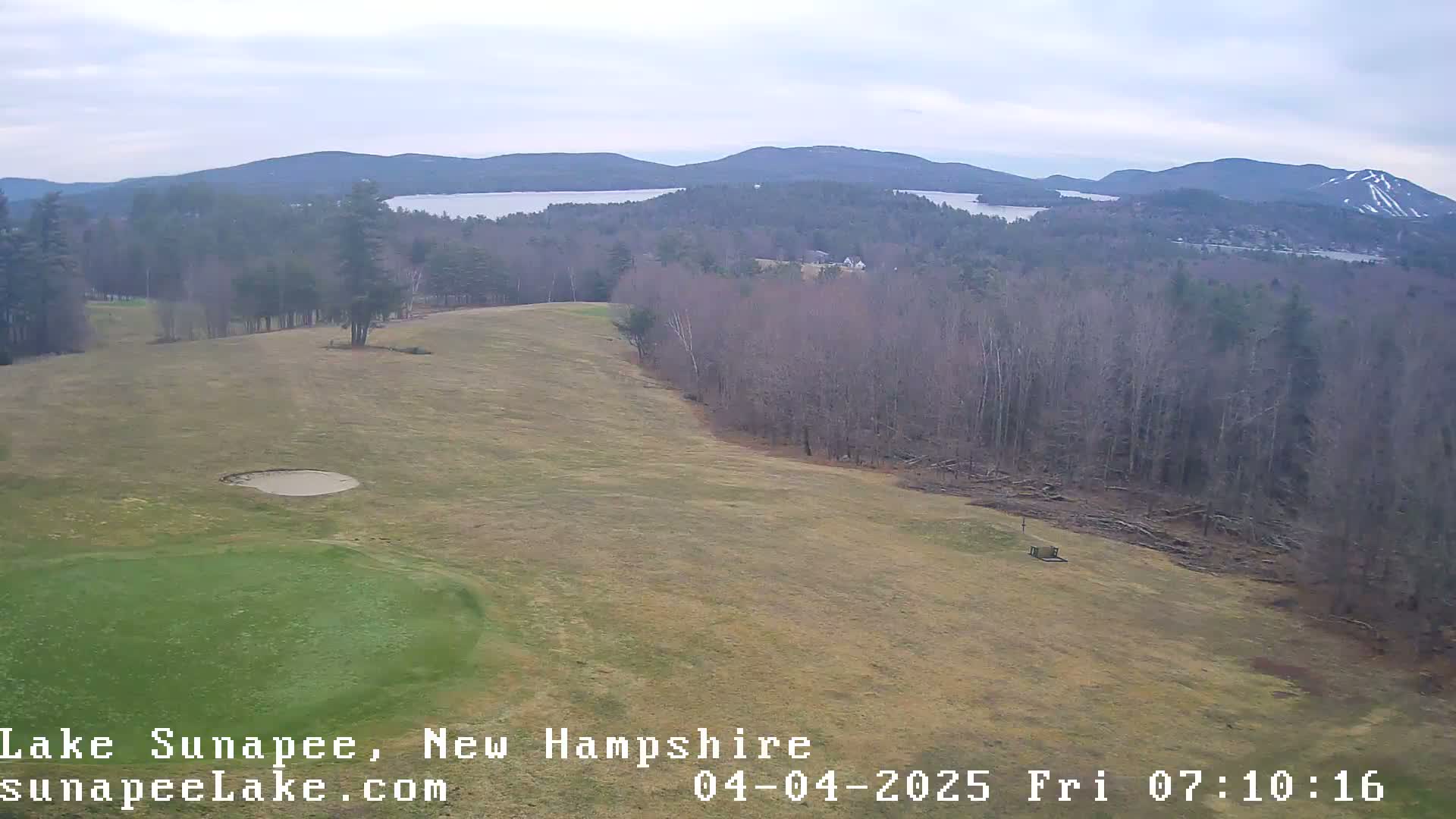 An elevated view of a golf course with a sand trap and a distant lake under an overcast sky.