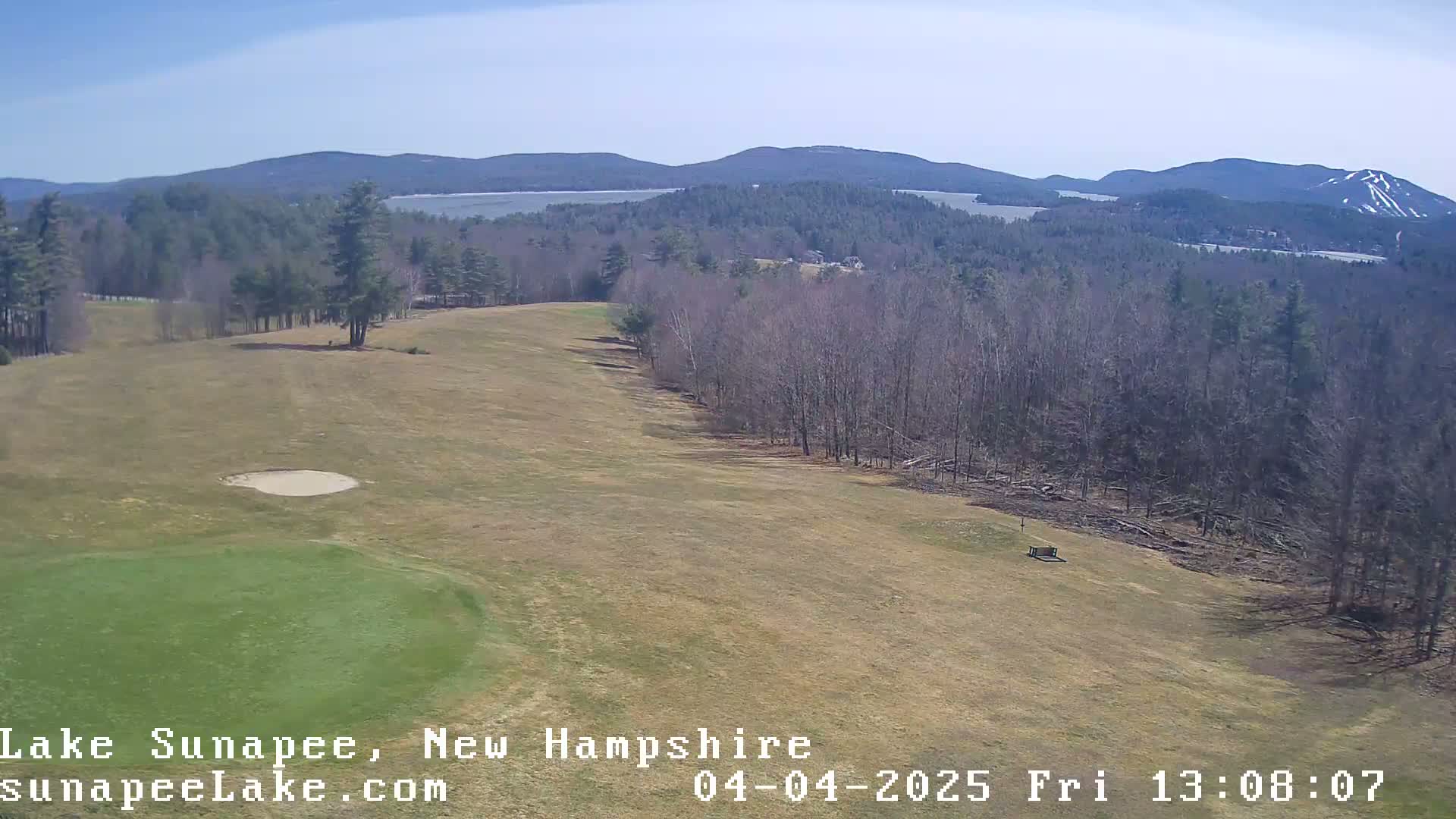 A high-angle view of a golf course fairway with a sand trap, bordered by leafless trees and overlooking a lake and distant mountains under a clear blue sky.
