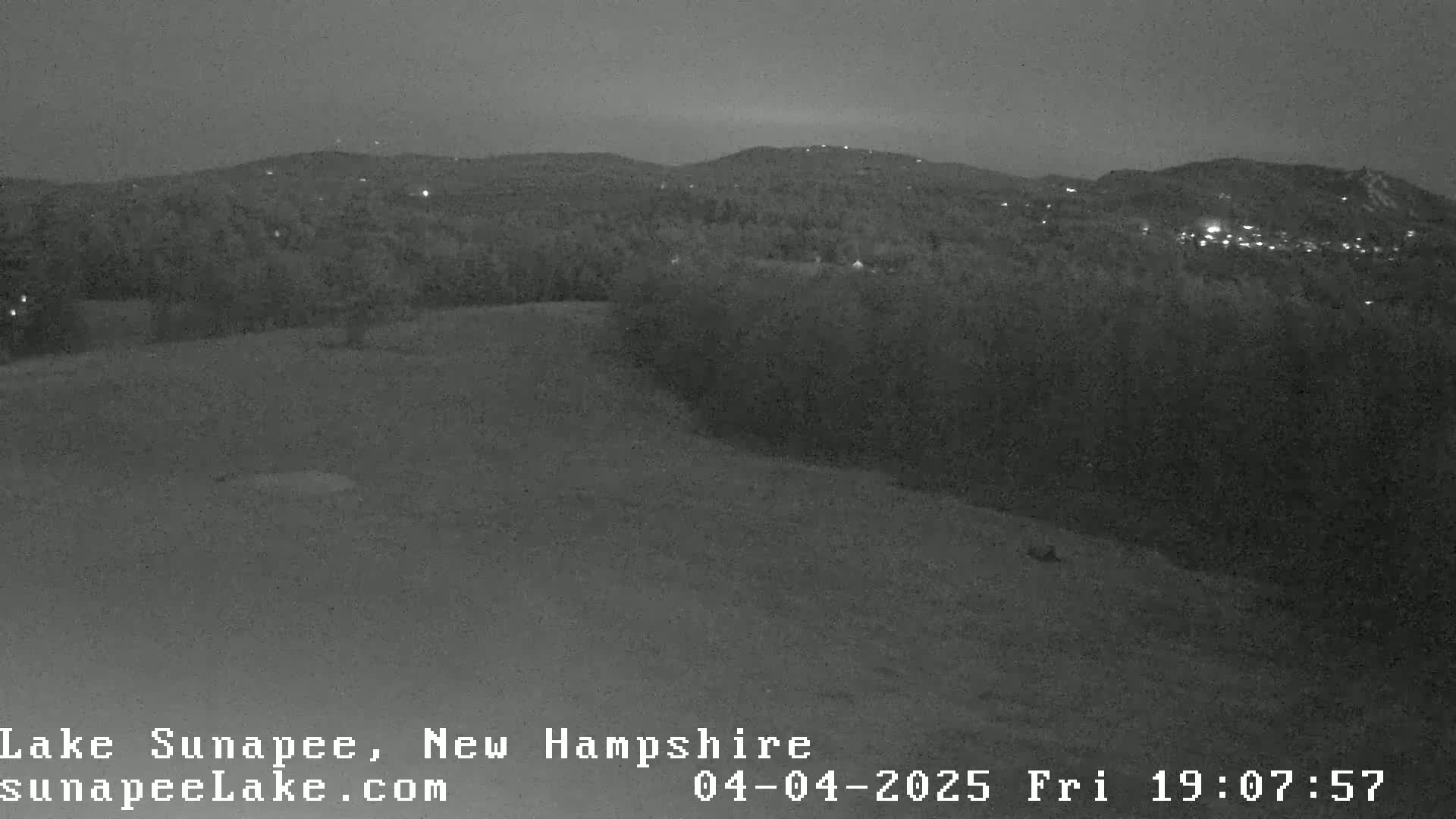 A nighttime, low-light view shows a dark, hilly landscape with sparse lights visible in the distance.