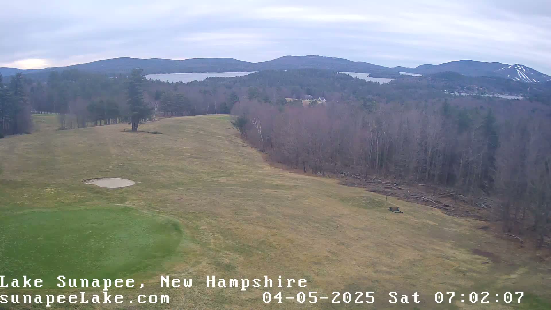 A mostly cloudy day overlooks a golf course green and fairway, with distant mountains and a lake visible in the background.
