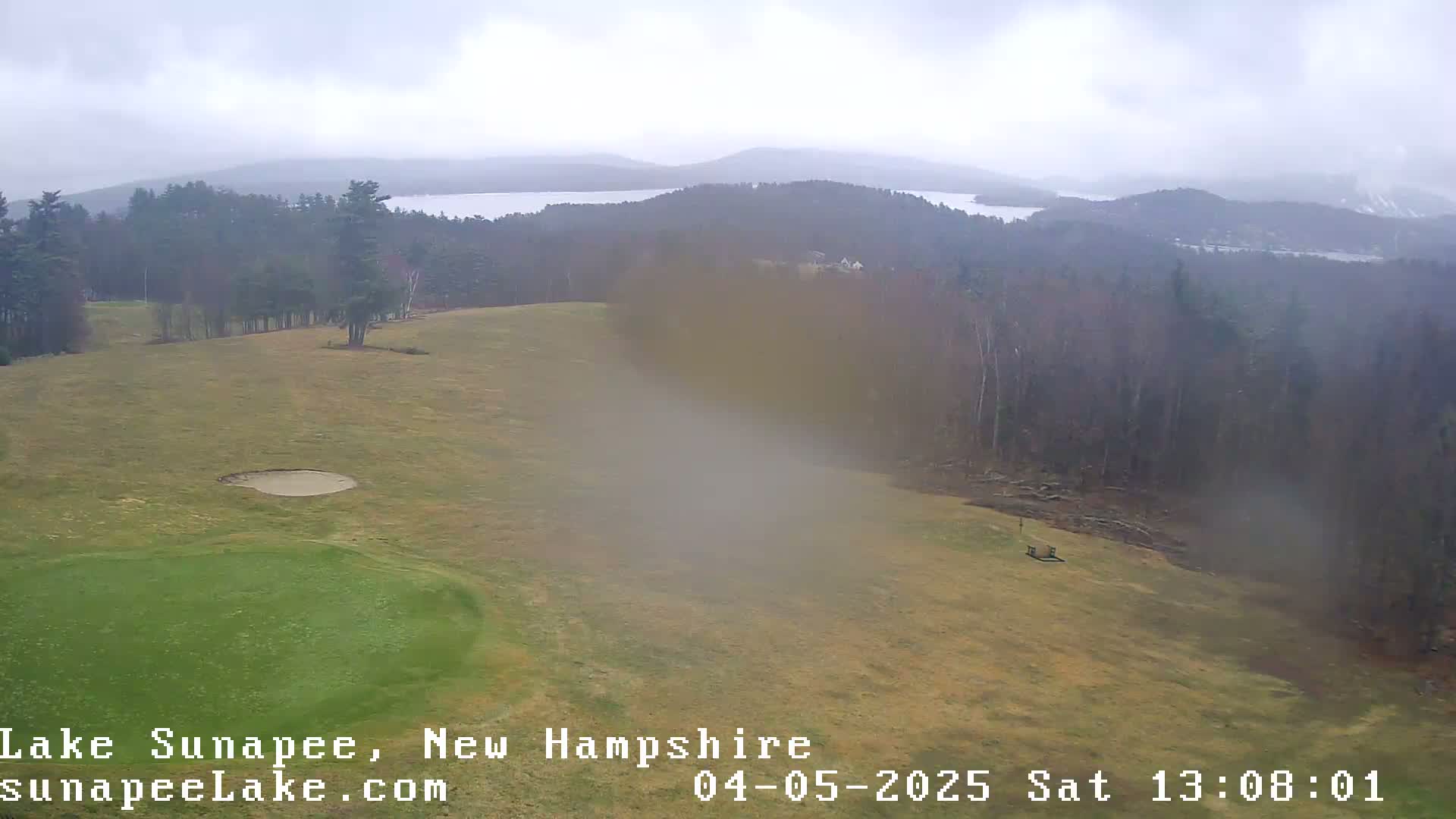 A high-angle view shows a golf course with a sand trap and green, overlooking a lake and distant, wooded hills under an overcast sky.