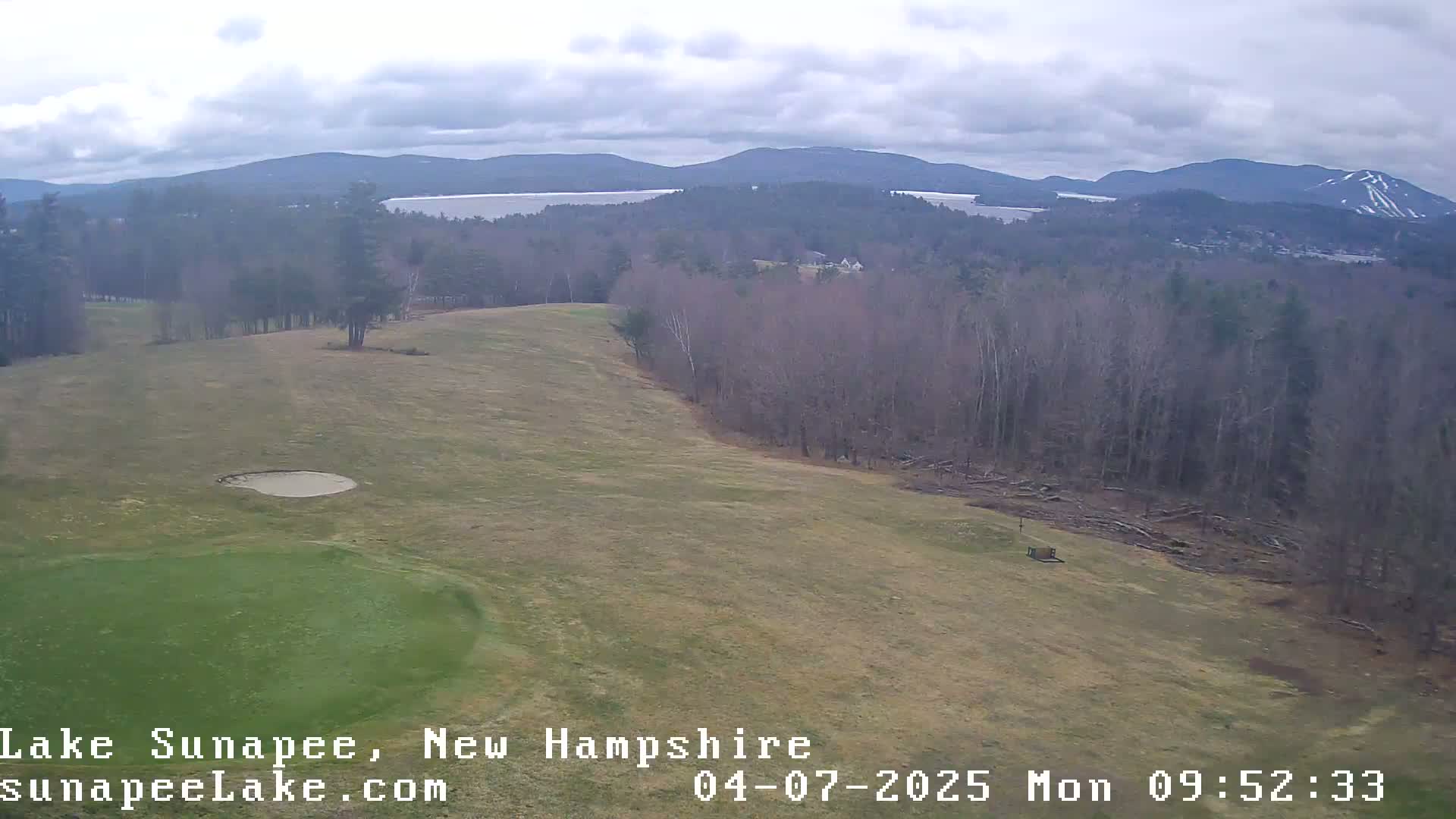 An elevated view of a golf course with a sand trap, a wooded area, and a lake and mountains in the distance under an overcast sky.