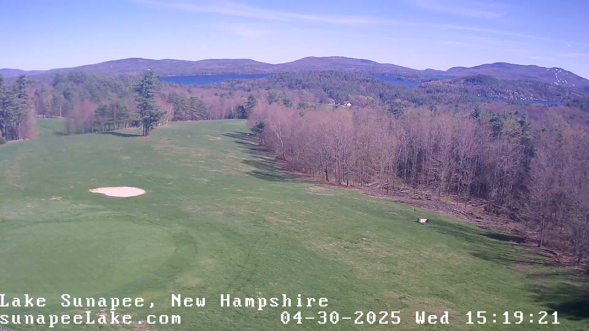 A sunny view of a golf course fairway with a sand trap, bordering a forest and overlooking a lake and distant mountains.