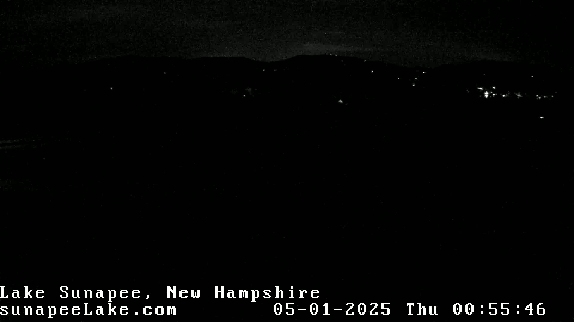 A nighttime, long-distance view of a dark landscape with a few scattered lights in the distance.
