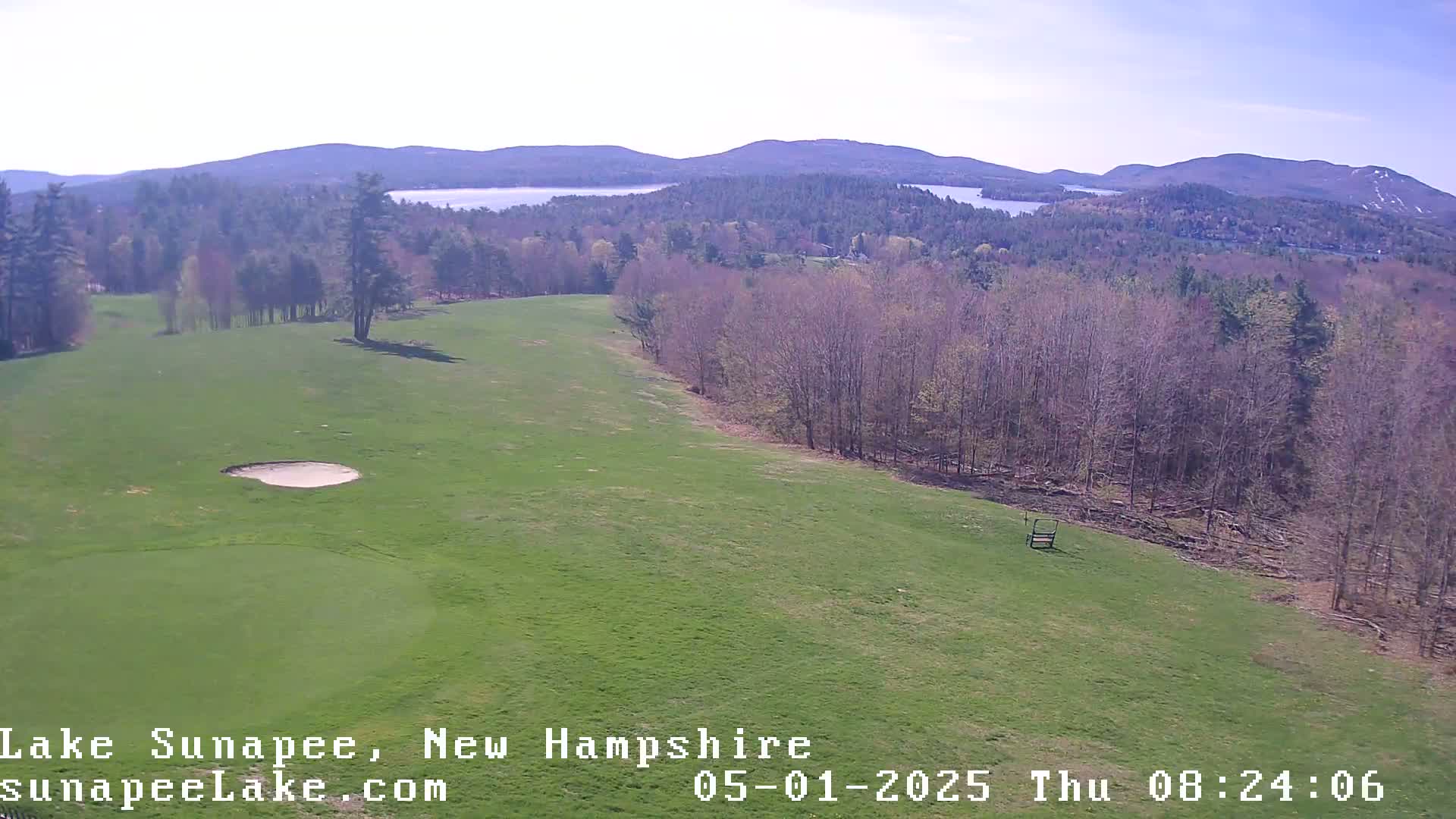 A high-angle view shows a green golf course with a sand trap, overlooking a forest and distant lakes under a clear sky.