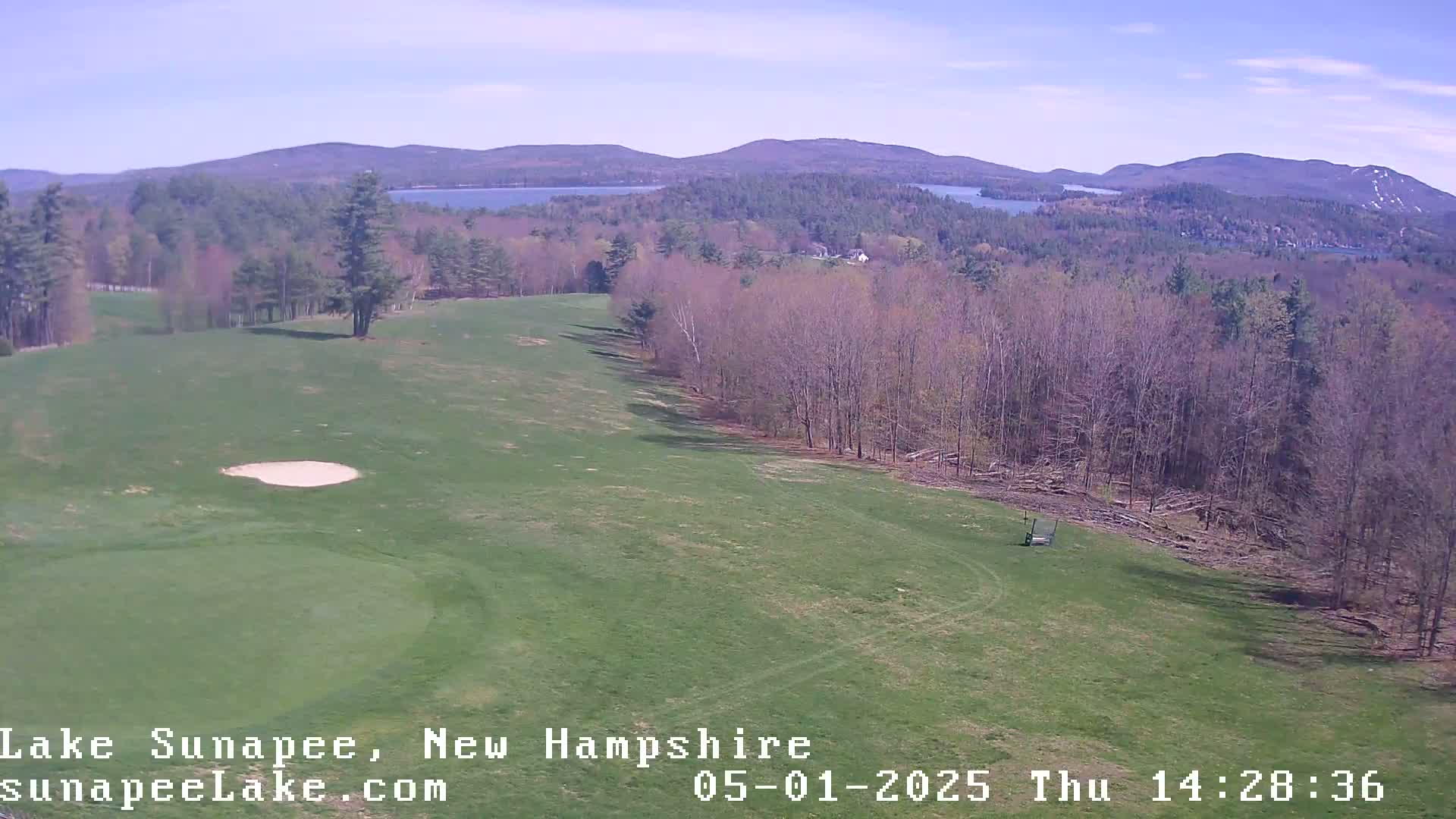 A golf course green with a sand trap, overlooking a lake and mountains under a mostly clear sky.