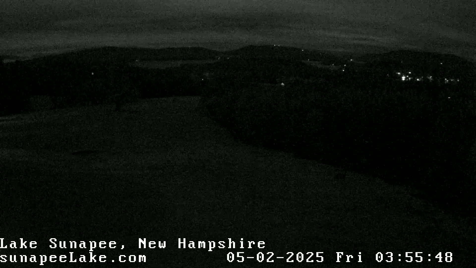 A dark, nighttime view of a hilly landscape with a few distant lights visible under a mostly clear sky.