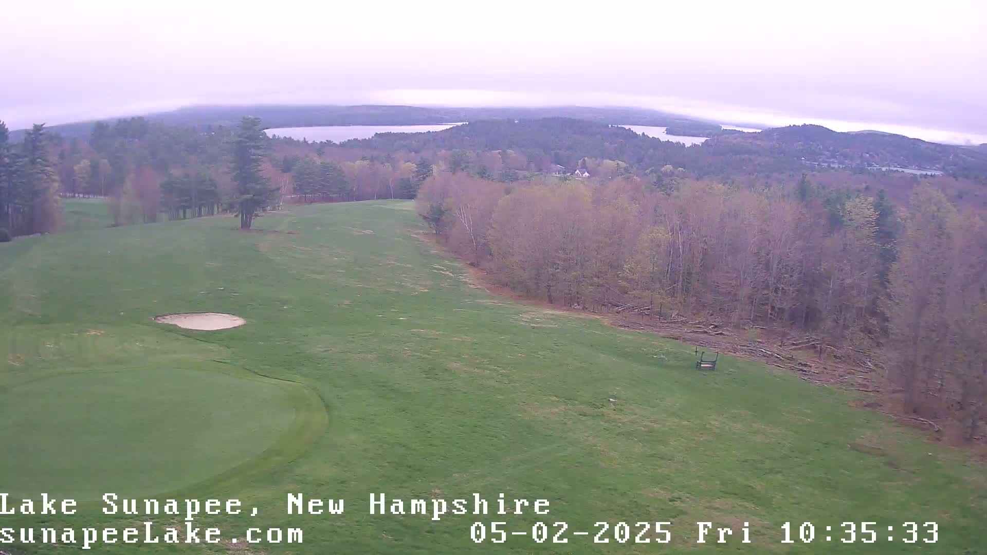A golf course with a sand trap, bordered by a wooded area overlooking a hazy lake in the distance.