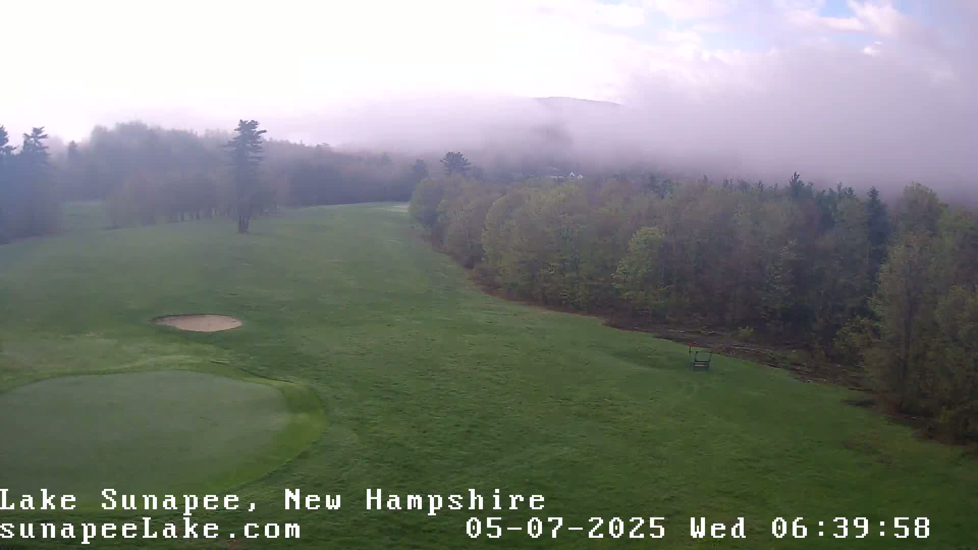 A foggy morning reveals a verdant golf course bordered by a line of trees and hills shrouded in mist.
