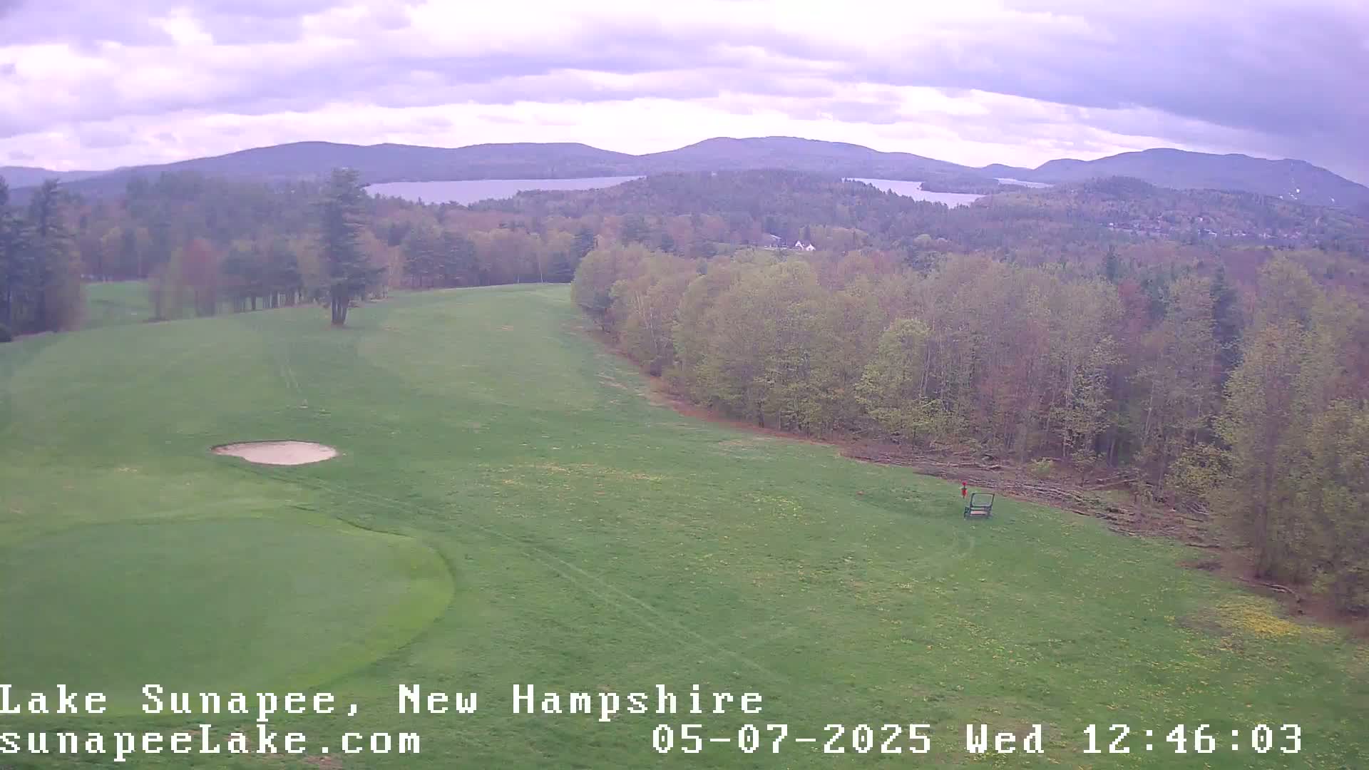 An aerial view shows a golf course with a sand trap, bordered by a forest and overlooking a lake and distant mountains under a partly cloudy sky.