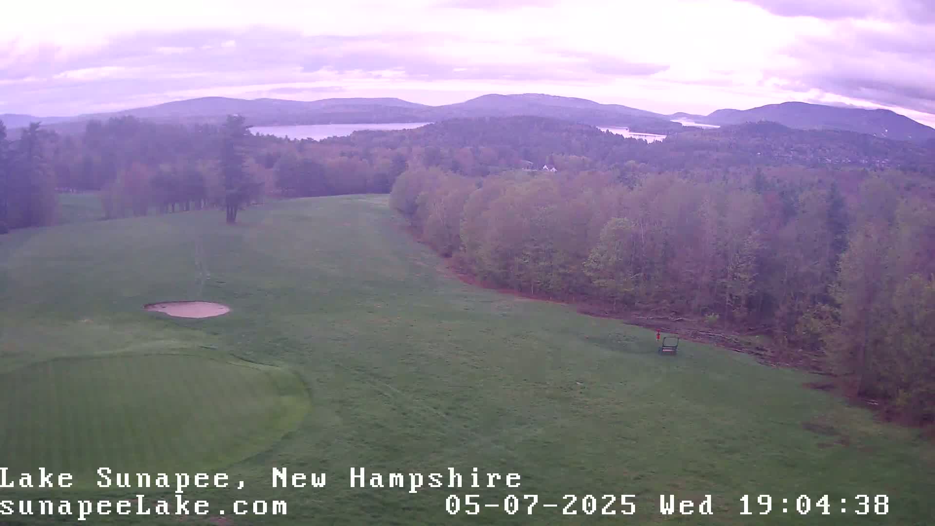 An aerial view shows a golf course with a sand trap and putting green, situated next to a forest with a lake and mountains visible in the distance under a cloudy, purplish sky.