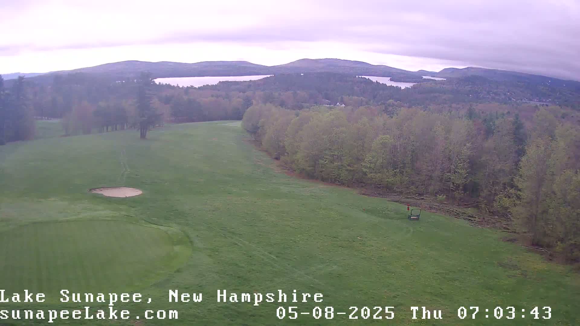 An aerial view shows a golf course with a sand trap and green, overlooking a lake and forested hills under an overcast sky.