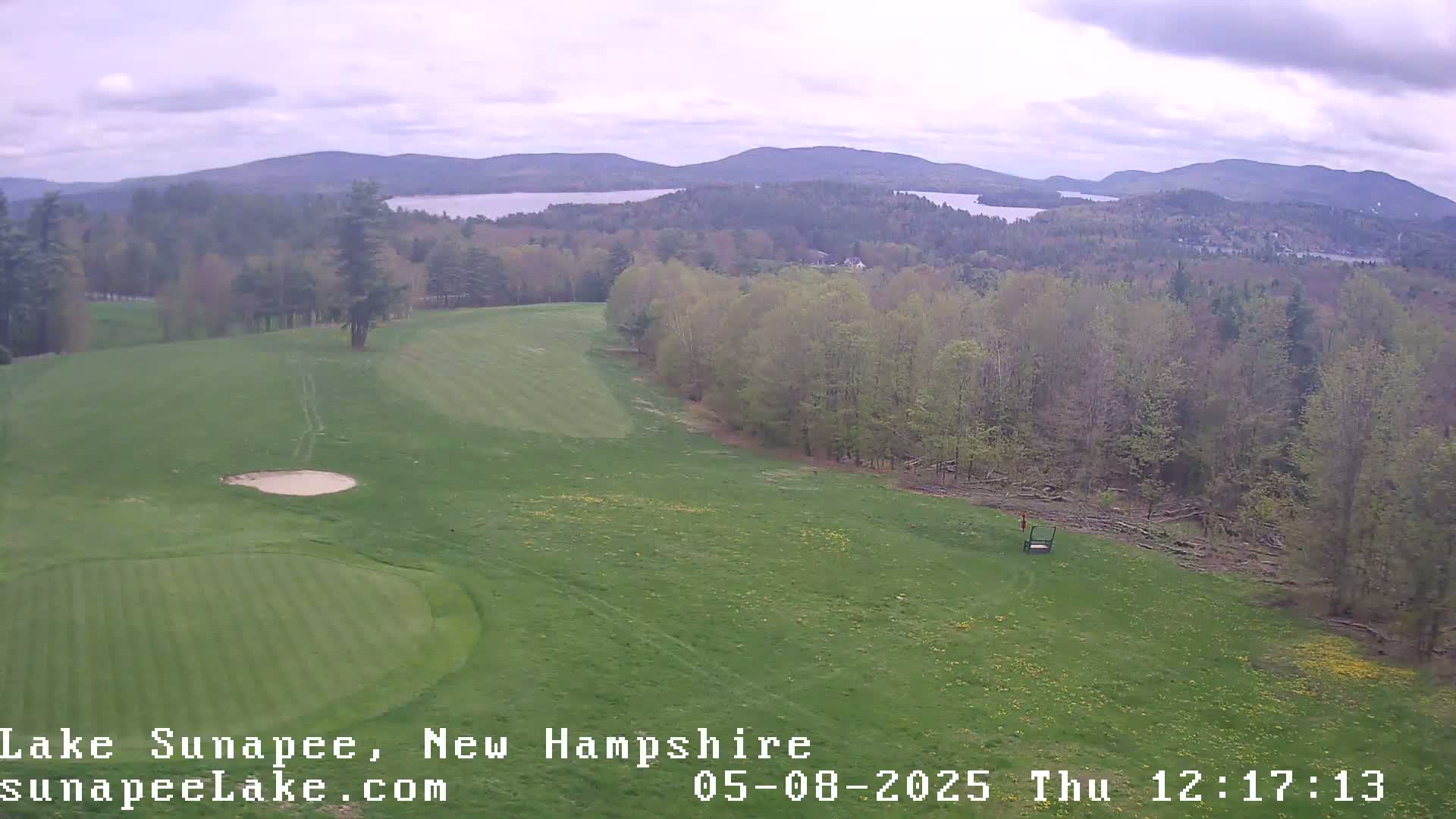 An aerial view shows a golf course with a green and sand trap, overlooking a lake and distant mountains under a cloudy sky.