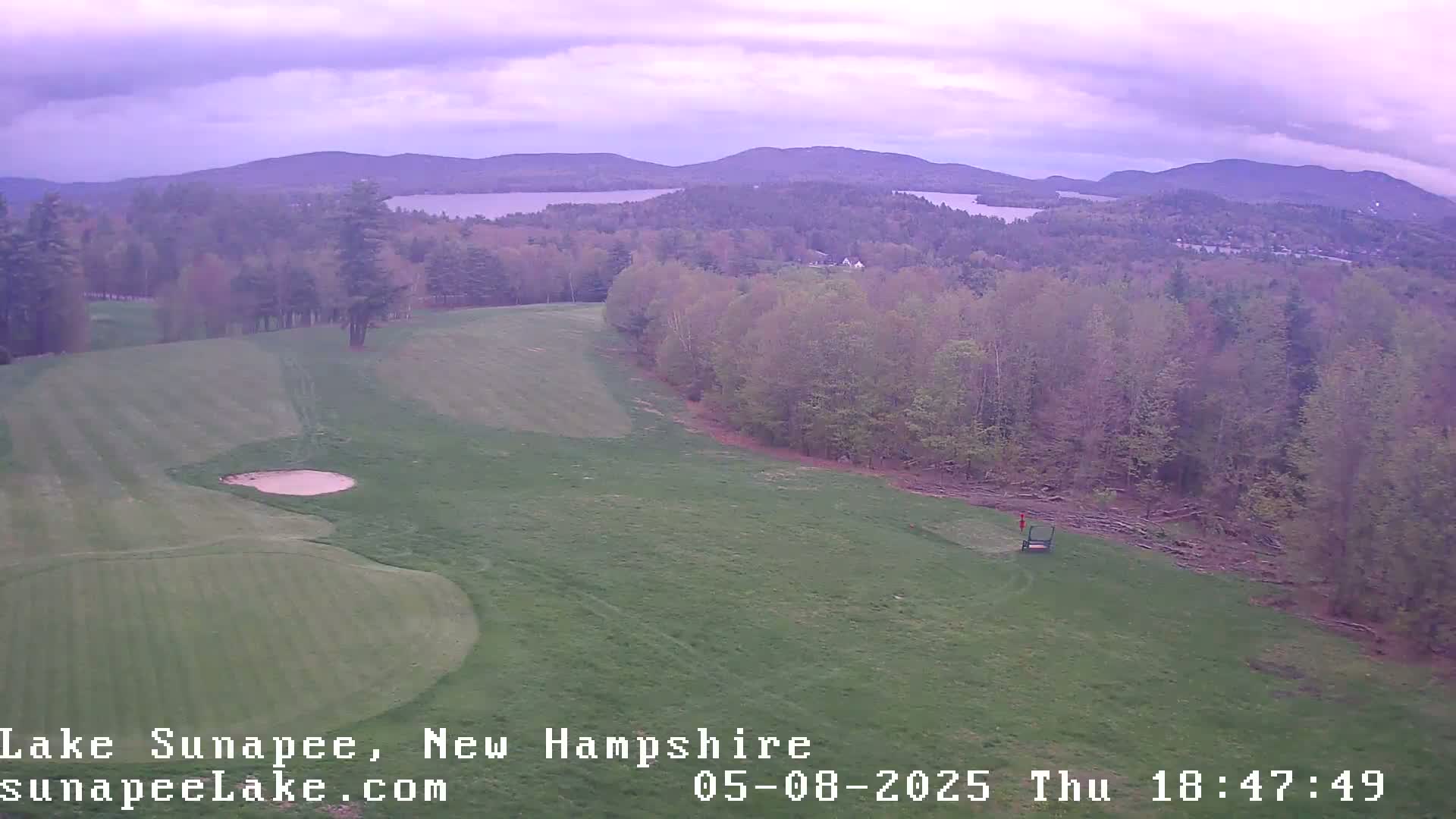 A golf course with a sand trap and distant lake and mountains under an overcast sky.