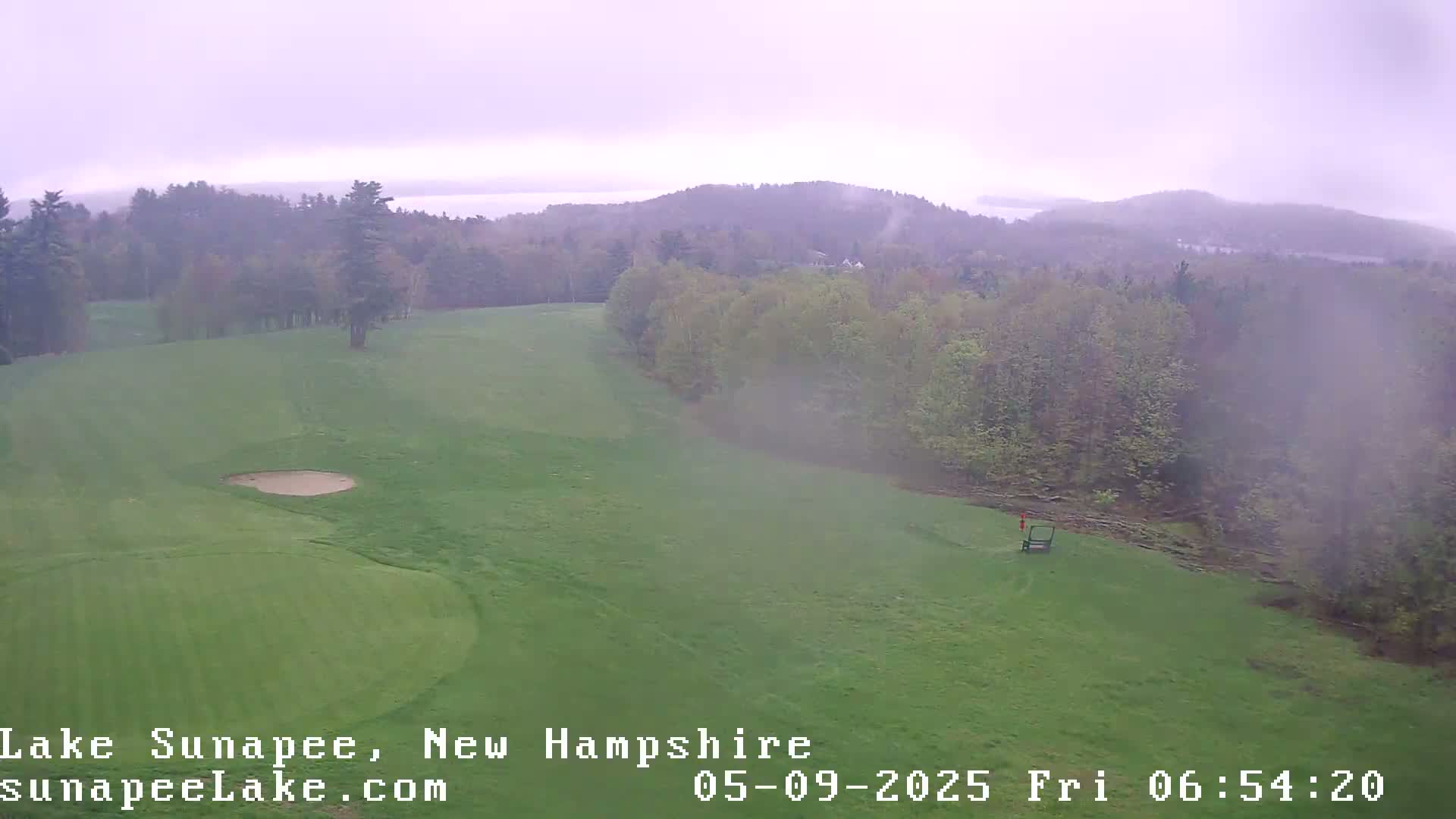A hazy, overcast view shows a golf course with a sand trap and a flag in the distance, overlooking a lake and forested hills.
