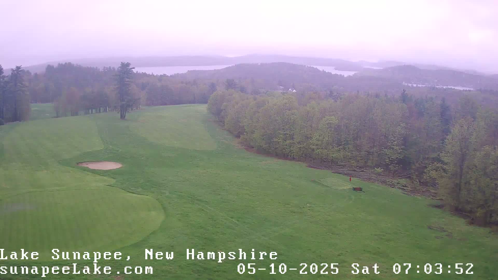 A hazy view shows a golf course with a sand trap, bordered by a forest overlooking distant lakes under an overcast sky.