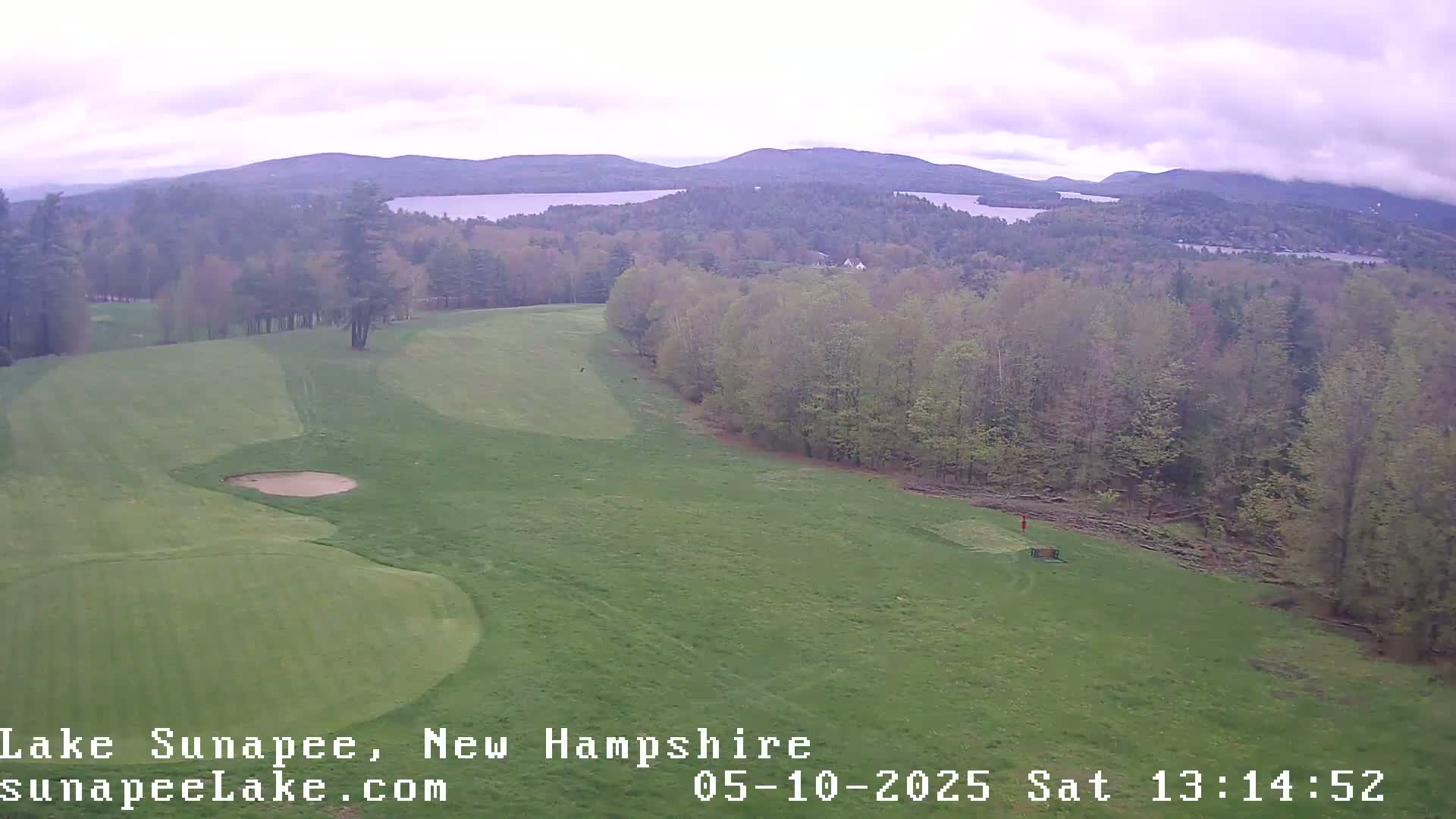 An aerial view shows a golf course with a sand trap, bordered by a forest overlooking a lake and distant mountains under an overcast sky.