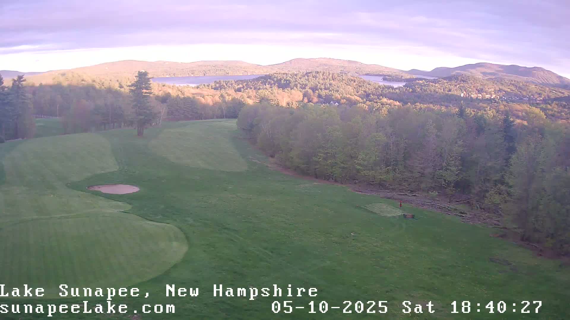 A golf course with a sand trap and a distant view of a lake and mountains under a soft, light sky.