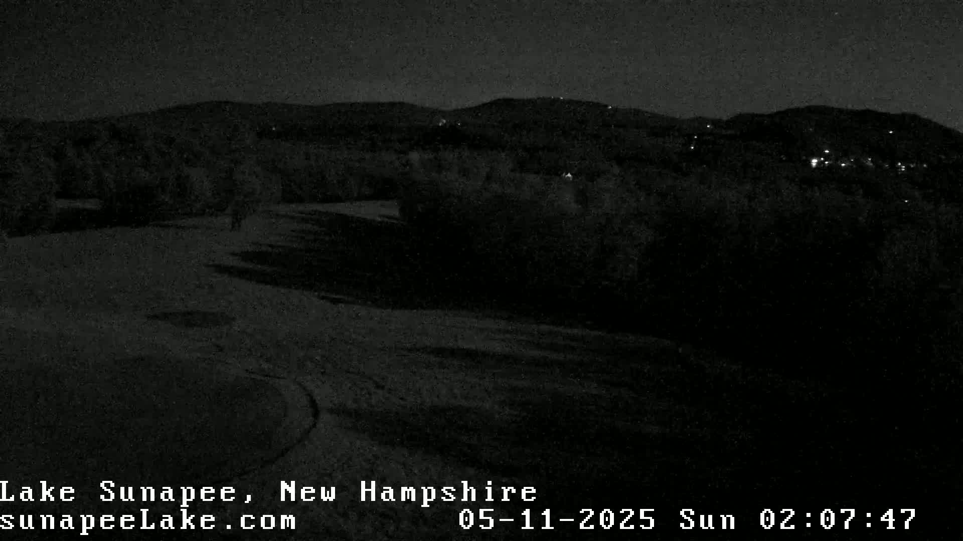 A nighttime, low-light view shows dark land and trees extending to distant, dimly lit hills under a dark sky.