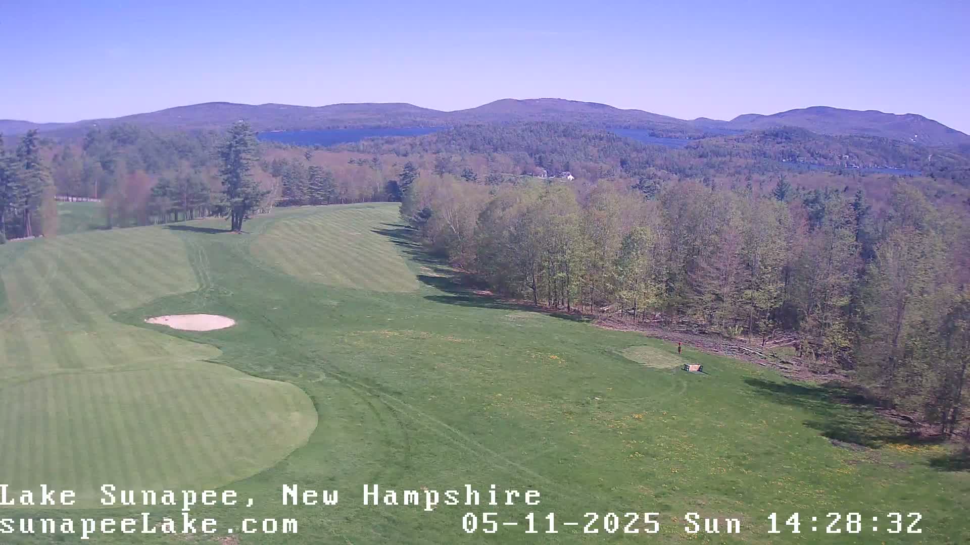 A sunny, expansive view of a golf course fairway with a sand trap, bordered by a line of trees and overlooking distant mountains and lakes.
